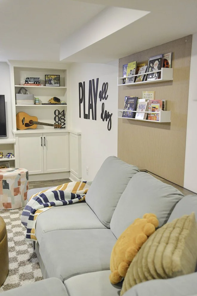 Cozy living room with a light gray sofa decorated with orange and tan pillows. A multicolored striped blanket is draped over the sofa arm. On the wall behind is a large black sign saying 'PLAY all day.' There are white wall-mounted shelves with books