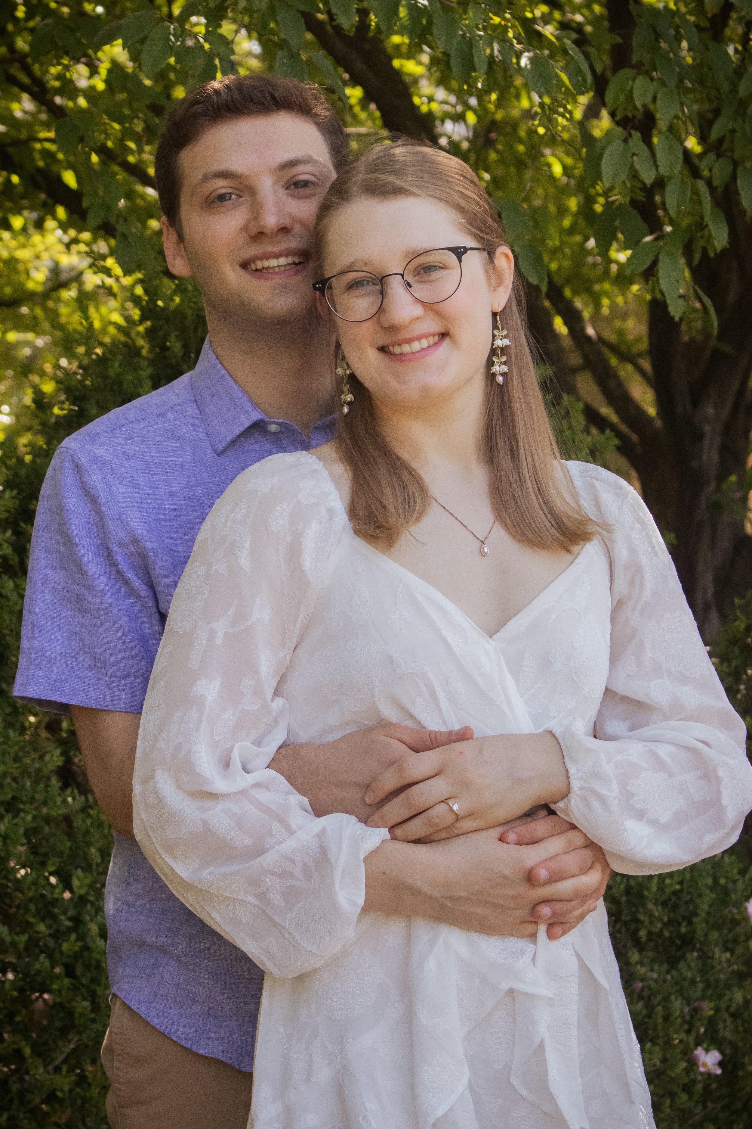 A young woman and man stand outdoors in front of green trees, smiling. The woman wears glasses, earrings, a white dress, and a necklace, and a wedding ring on her left hand. The man stands behind her, hugging her with both arms, wearing a purple shir