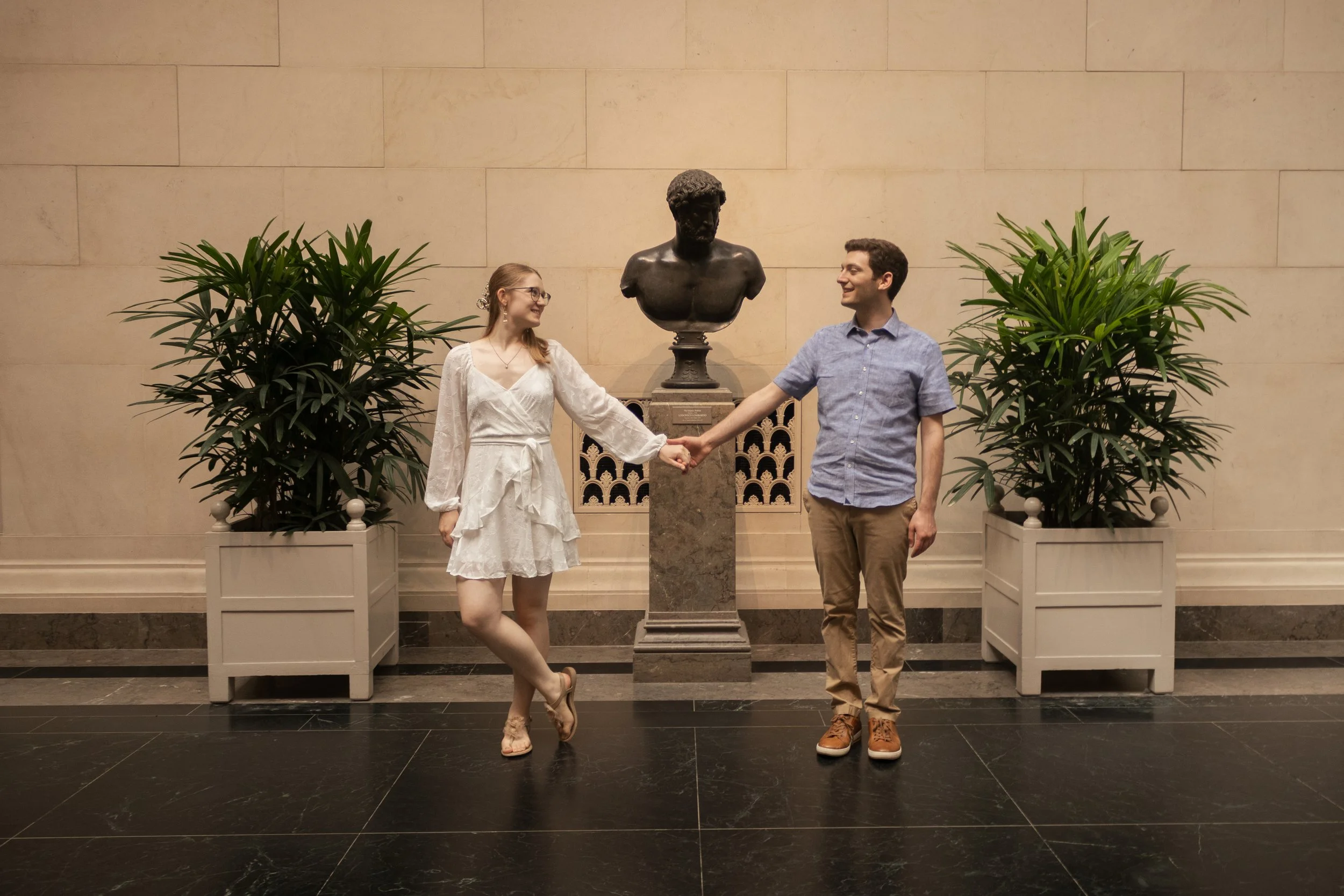 A man and woman holding hands in front of a bust sculpture inside a building with stone walls and black marble flooring.