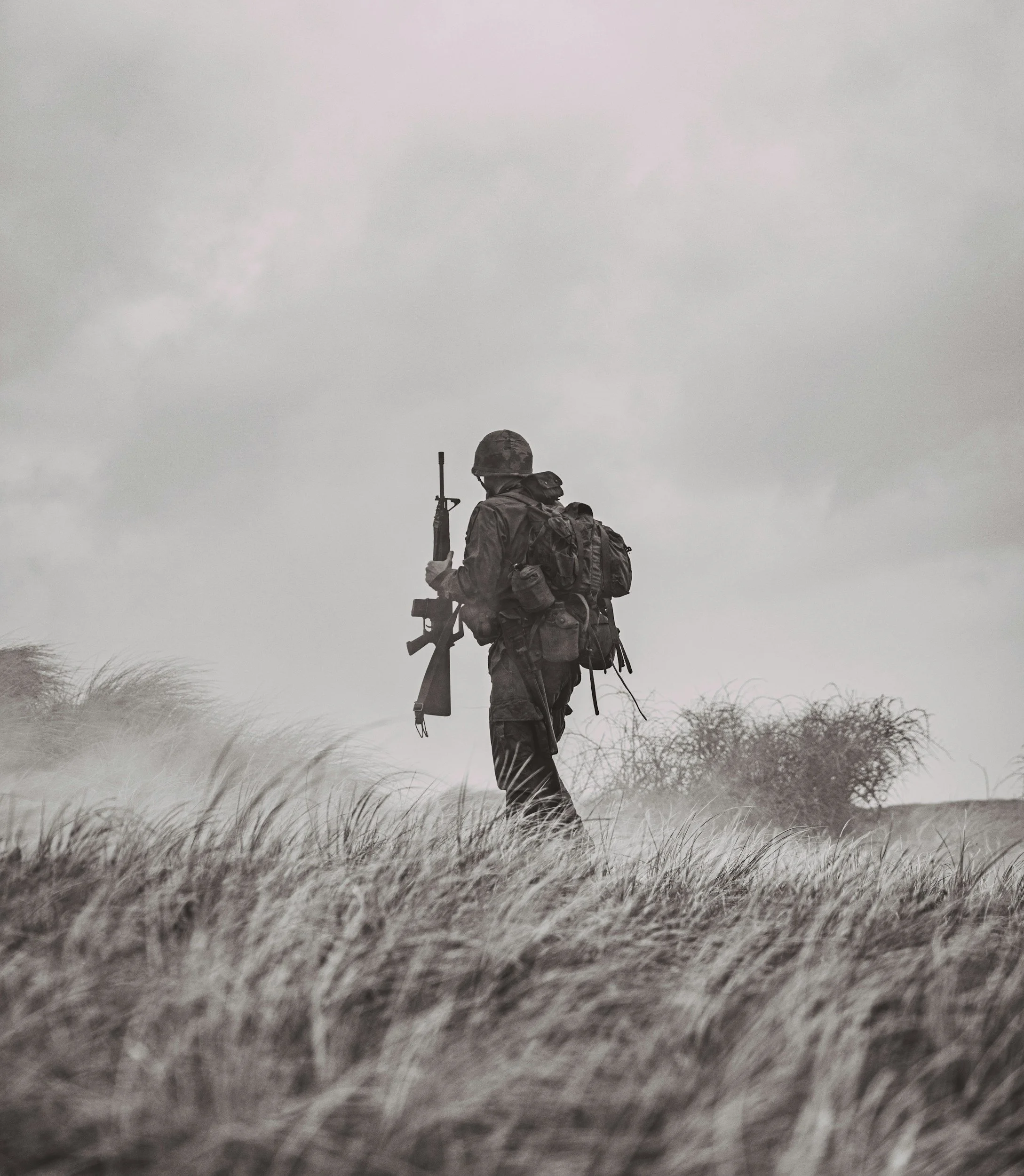 A soldier walking through a grassy field, carrying a rifle and wearing a helmet and tactical gear, in black and white.