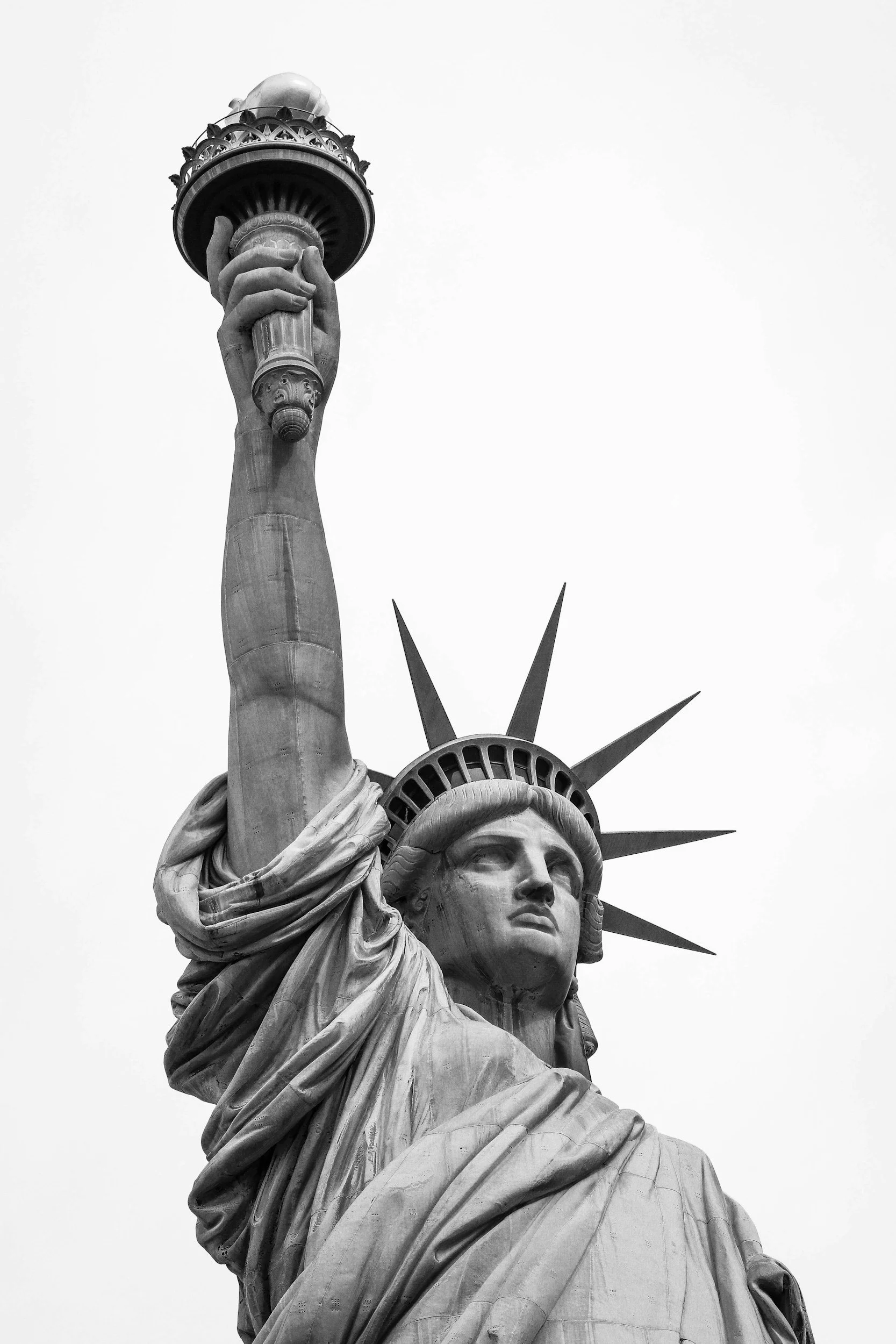 Close-up of the Statue of Liberty in black and white, showing the face, crown, and raised torch, with a plain sky background.