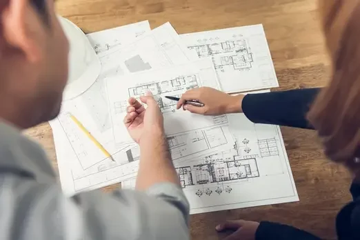 Two architects or engineers discussing architectural blueprints on a wooden table, with various detailed building plans and a yellow pencil.