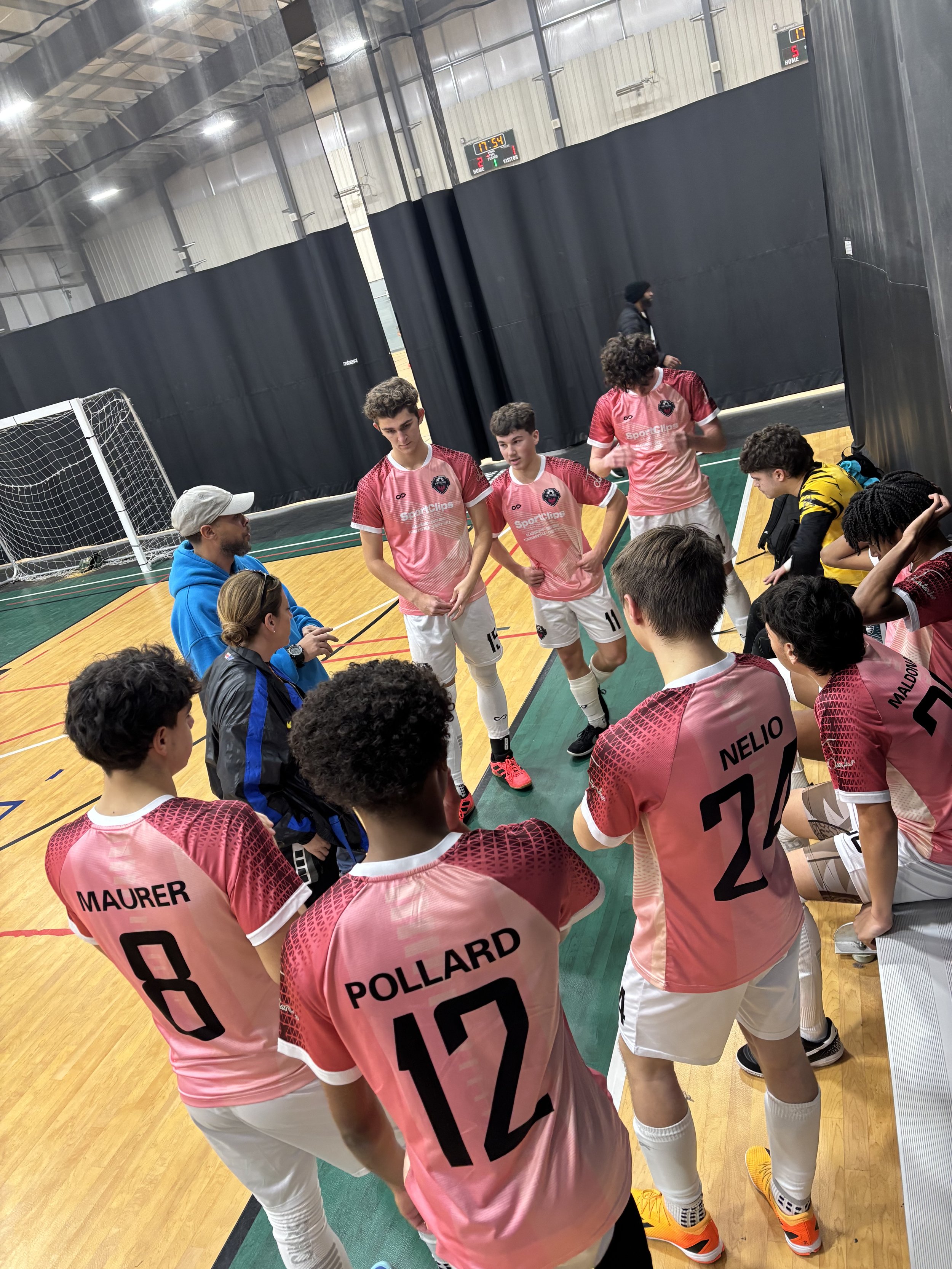 A group of young male soccer players wearing pink jerseys with black names and numbers, gathering around their coach in an indoor sports facility. The coach appears to be giving instructions while the players listen attentively. There is a goalpost i