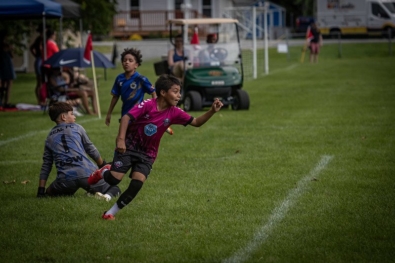 Young boys playing soccer on a grassy field, one boy in a pink jersey running past a goalie in gray, with others watching in the background.