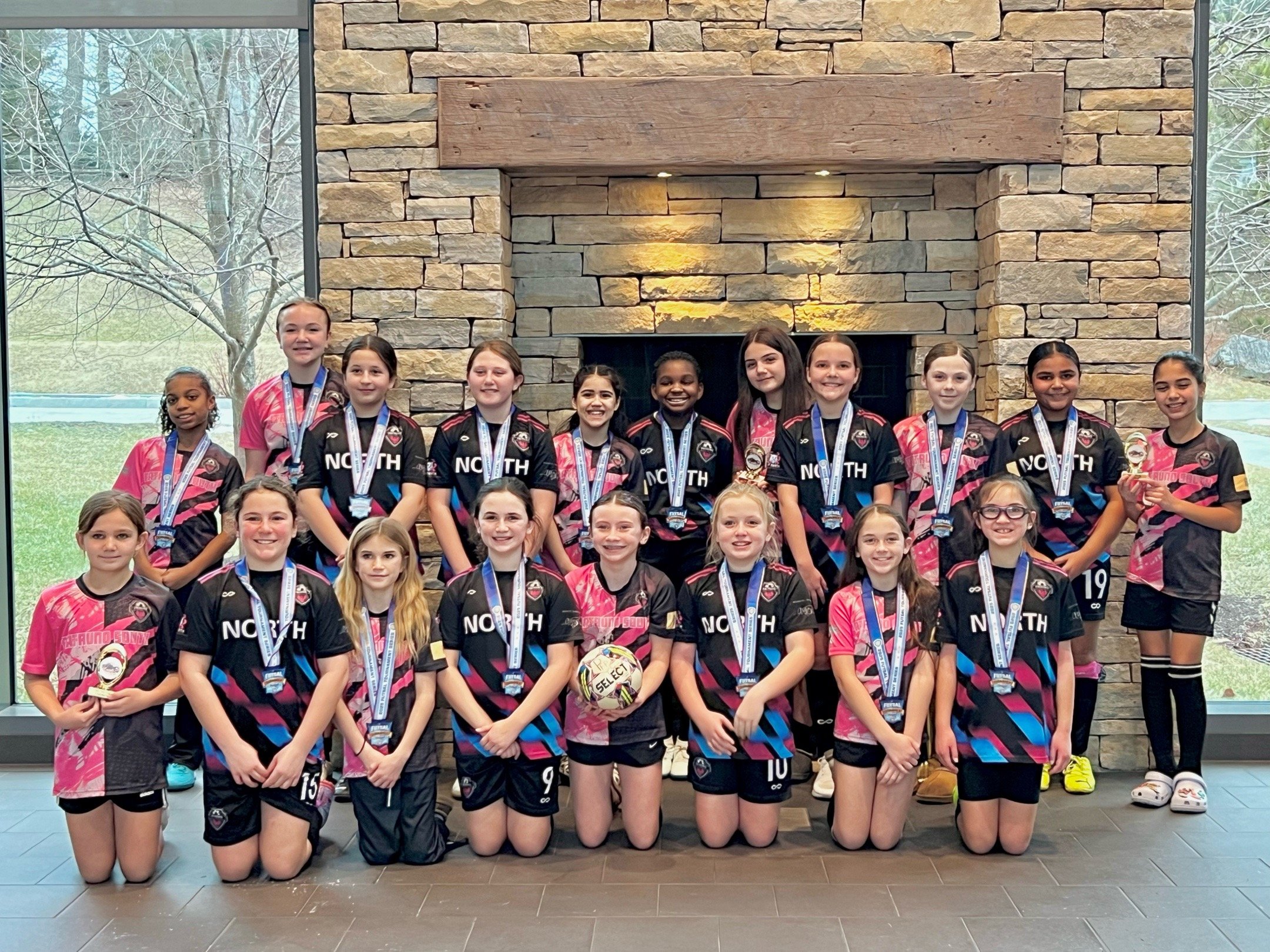 A group of young girls in sports uniforms wearing medals, posing indoors in front of a stone fireplace with large windows showing an outdoor landscape, celebrating a sports achievement.