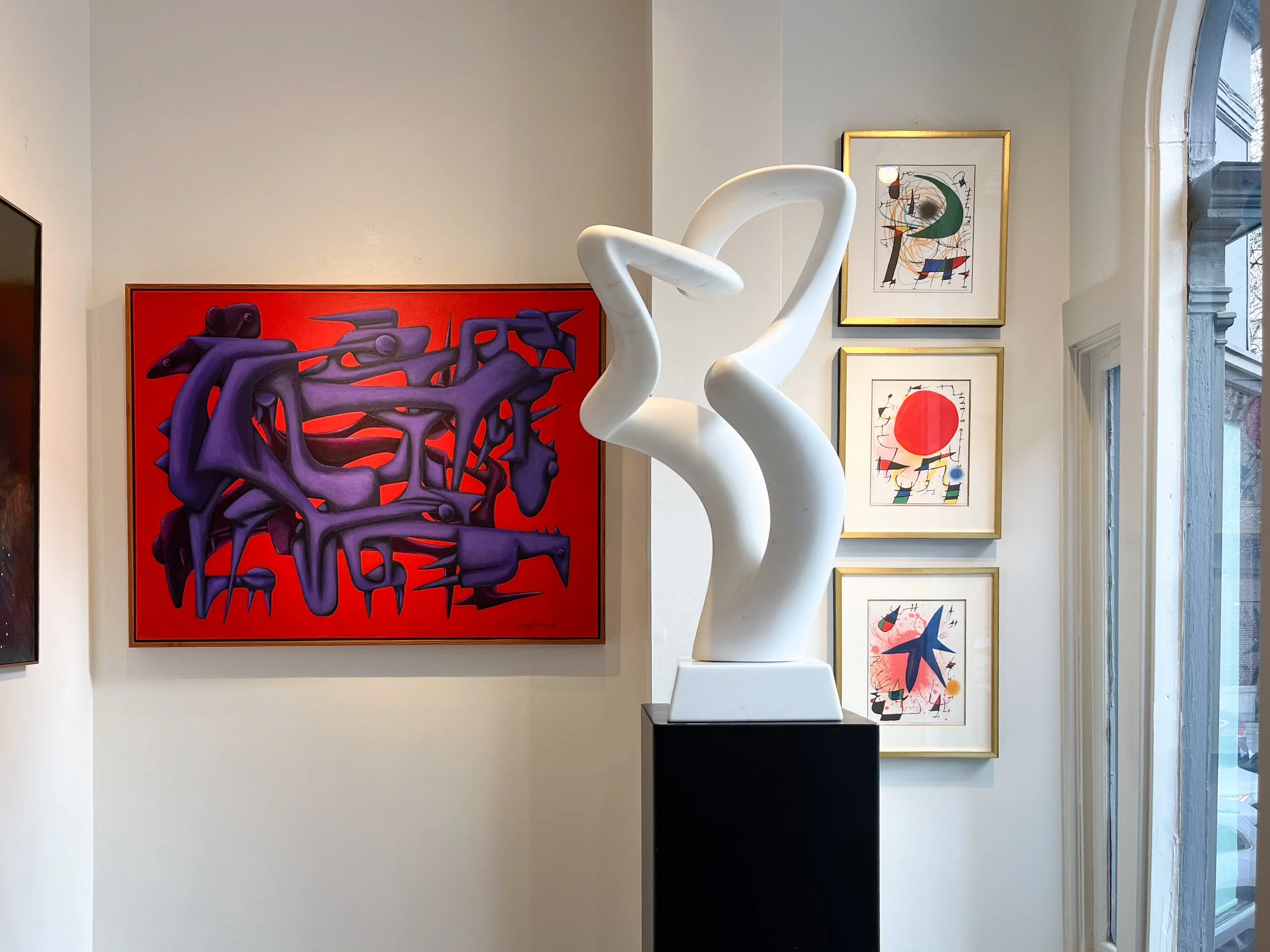 Interior photo of the gallery alcove, featuring a bright abstract red and purple painting, and abstract white marble sculpture, and three Miro lithographs. 