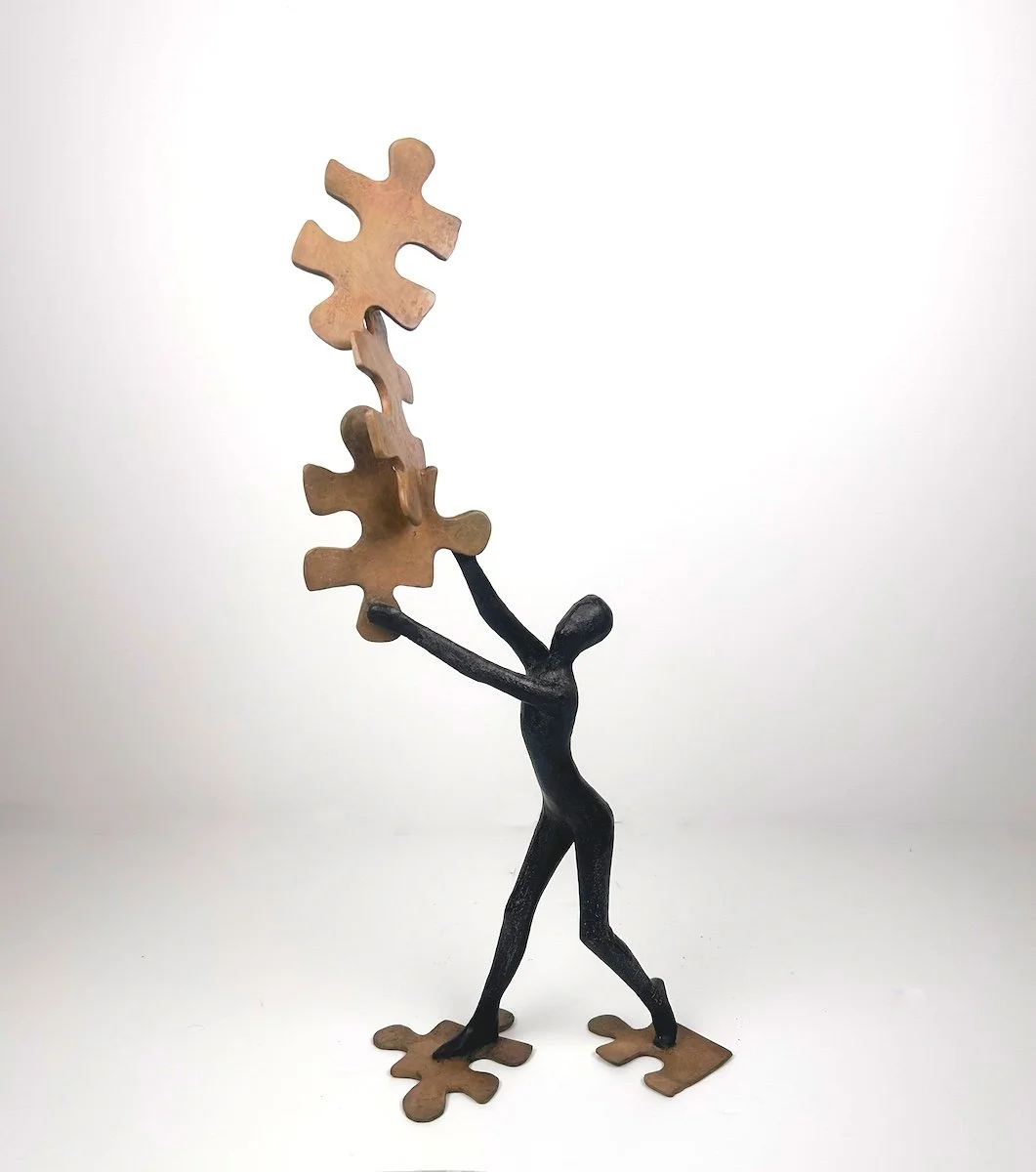 Tolla Puzzled, bronze figure standing on puzzle pieces and balancing several more above head, neutral colors