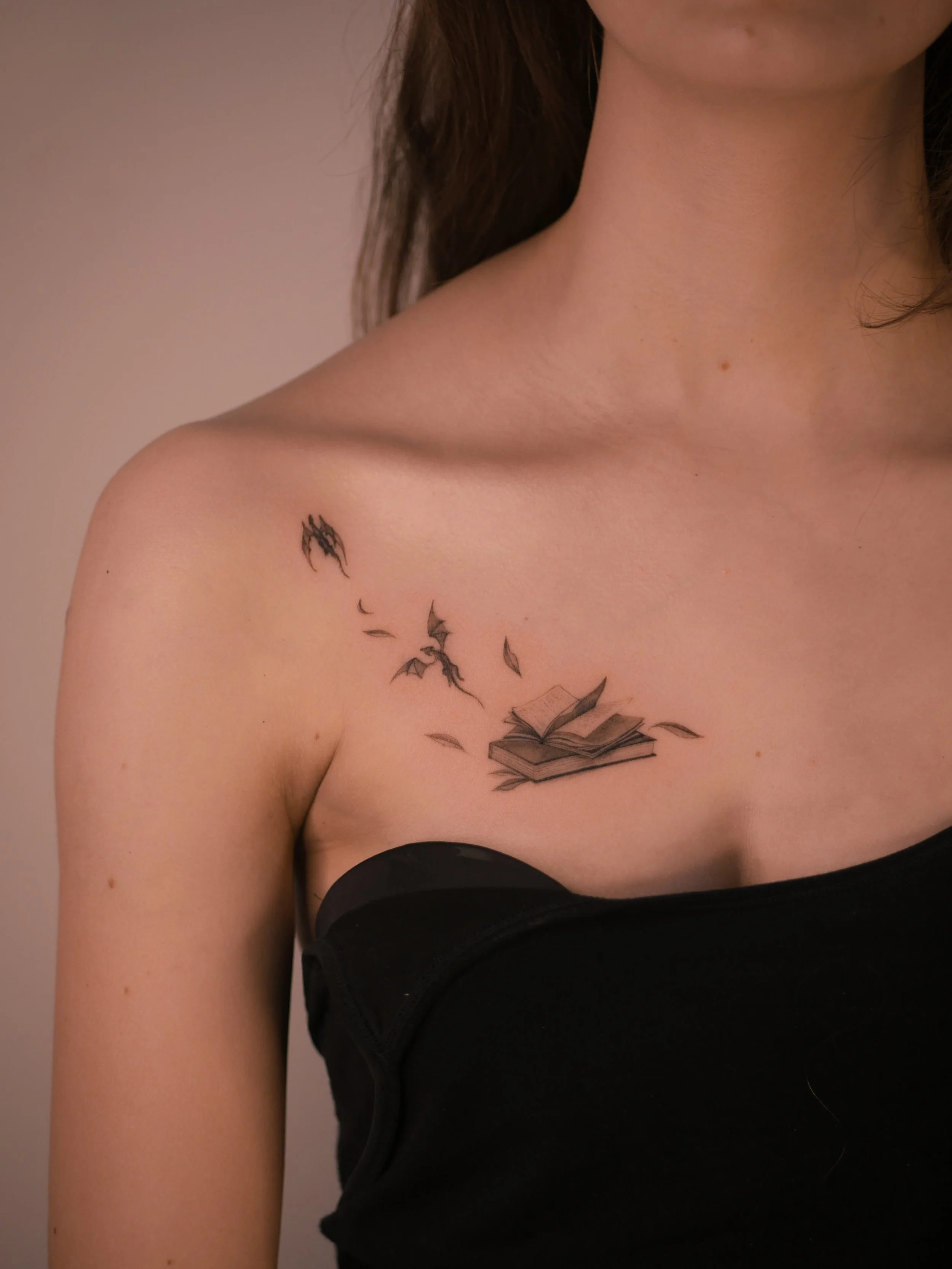 Fine-Line Tattoo eines offenen Buches mit fliegenden Seiten und Drachen, filigran gestochen am Schlüsselbein