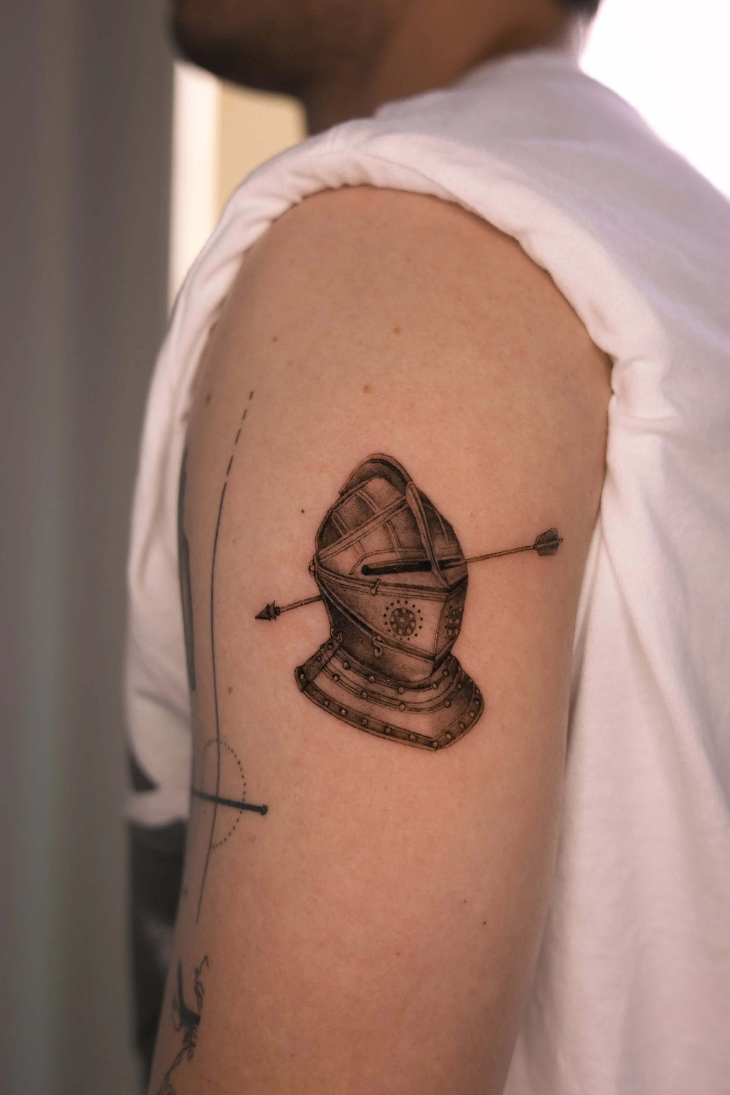 Fine line micro realism knight helmet tattoo with arrow detail on upper arm, black and grey delicate tattoo by Vienna tattoo artist