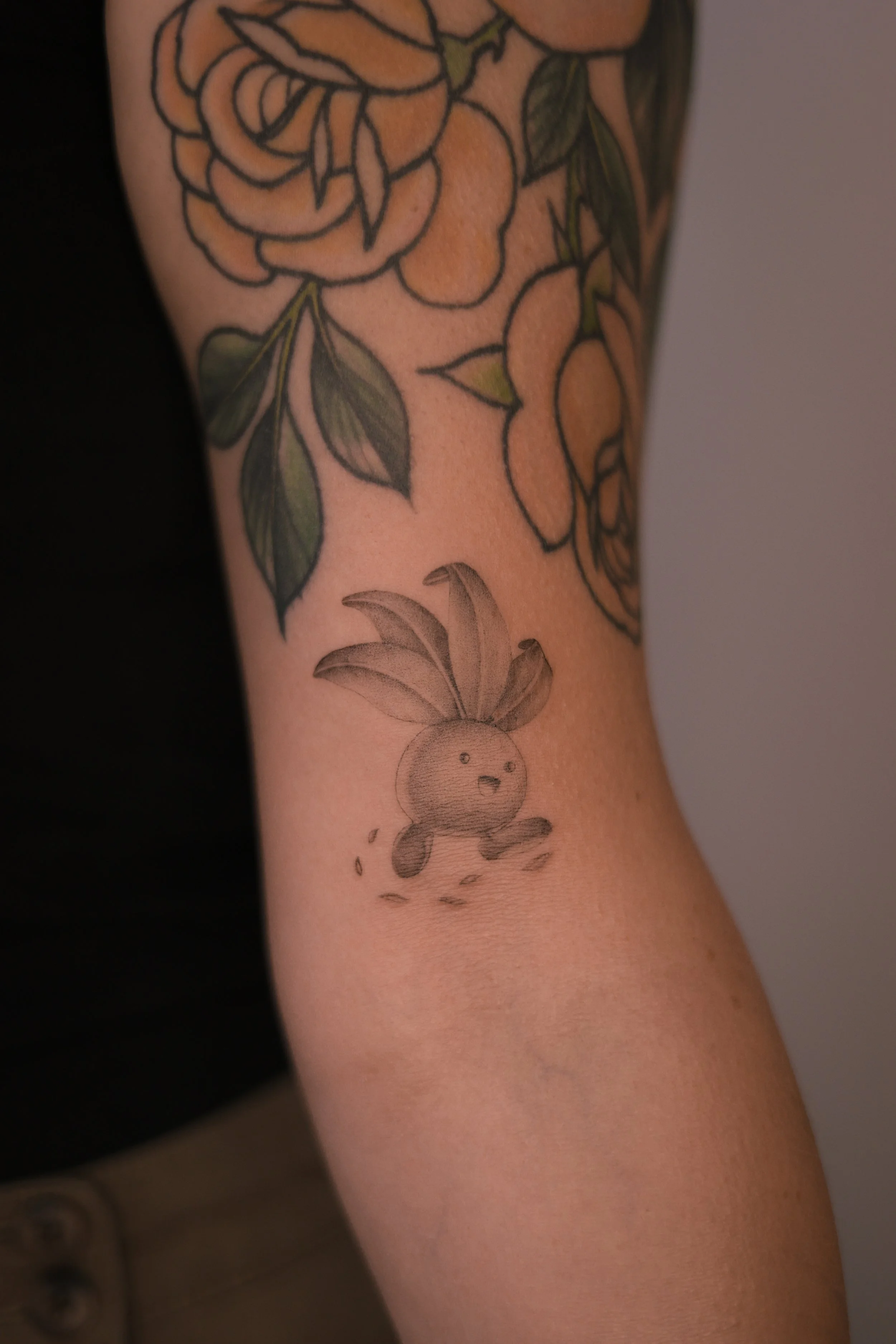Small black and grey Oddish Pokémon tattoo on the arm, with shaded leaves and a smiling face beneath existing floral tattoos, by a Vienna tattoo artist.