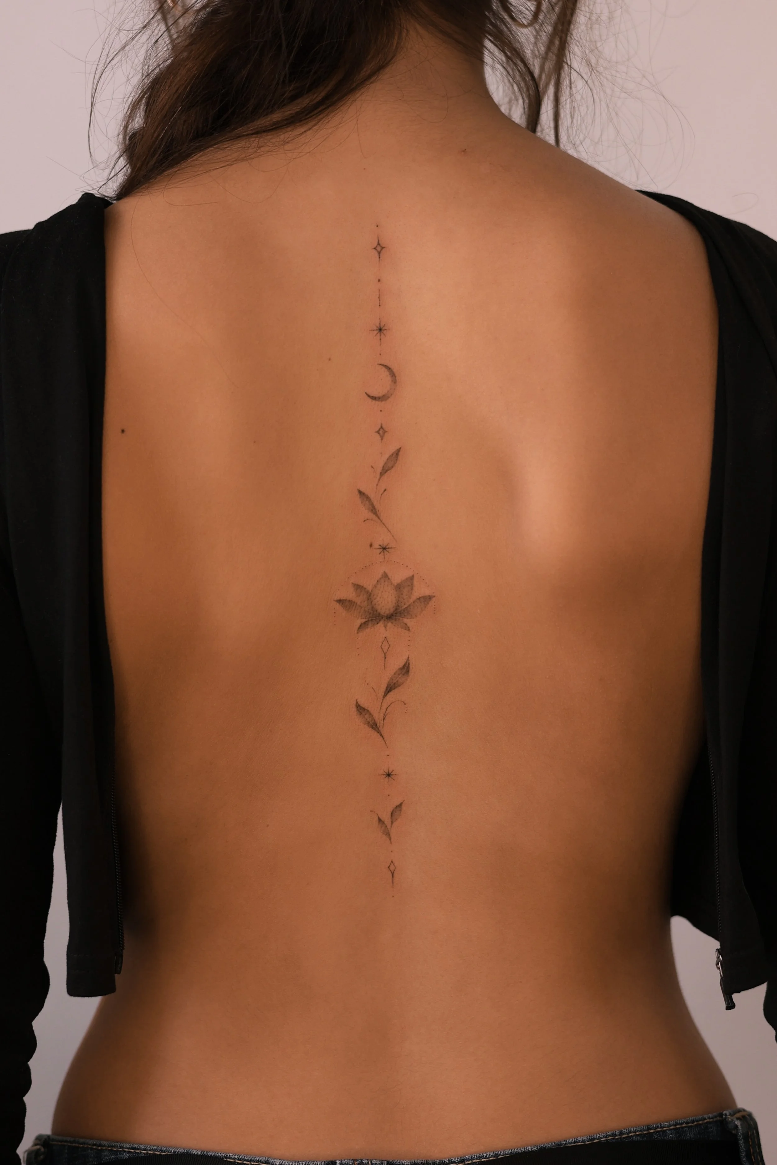 Fine line spine tattoo featuring a shaded lotus flower, crescent moon, stars and delicate leafy sprigs, by a Vienna tattoo artist.