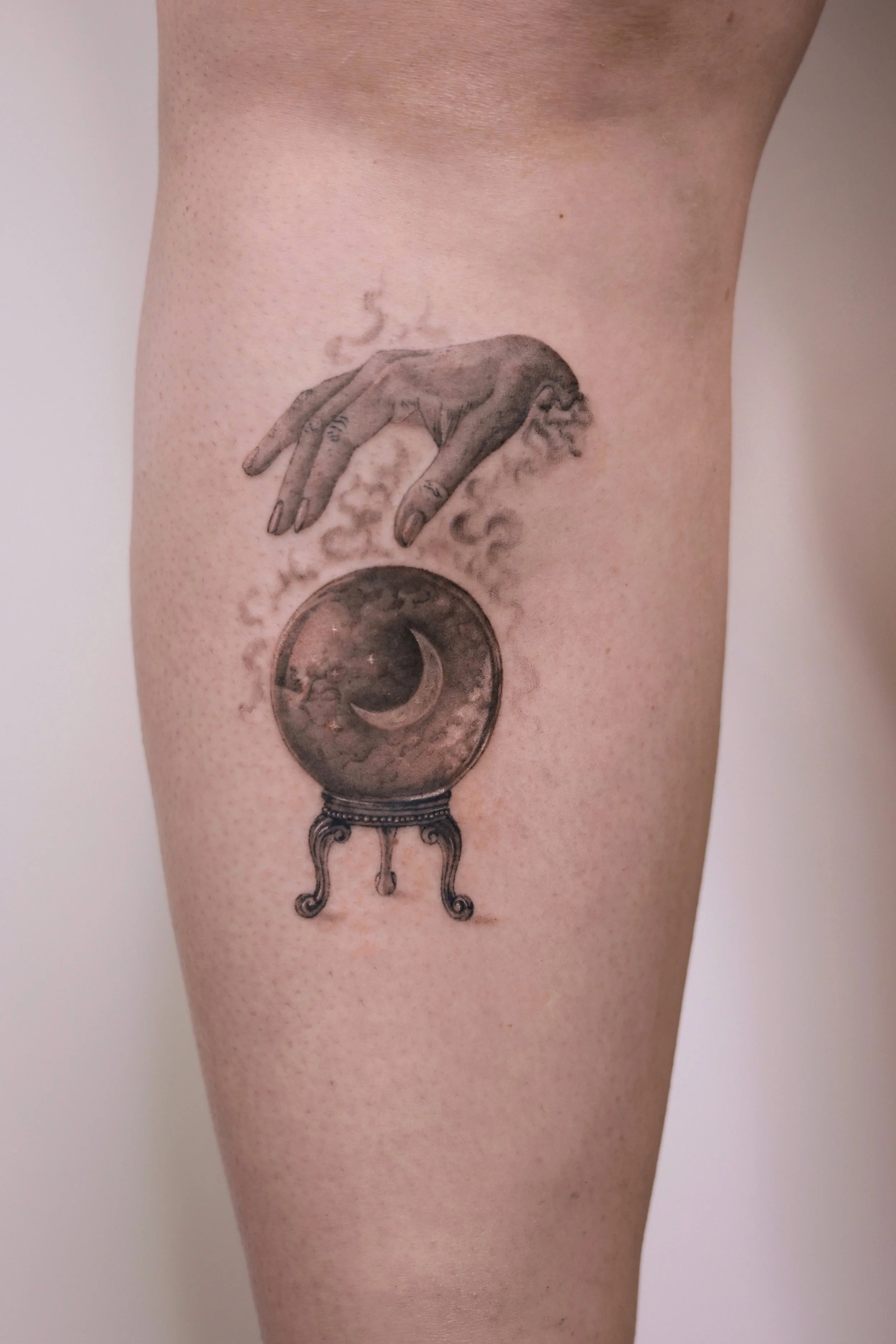 Hand über Kristallkugel mit Mondsichel – Mikro-Realismus/Fineline-Tattoo an der Wade; weiche Schatten, mystische Stimmung – Wien.