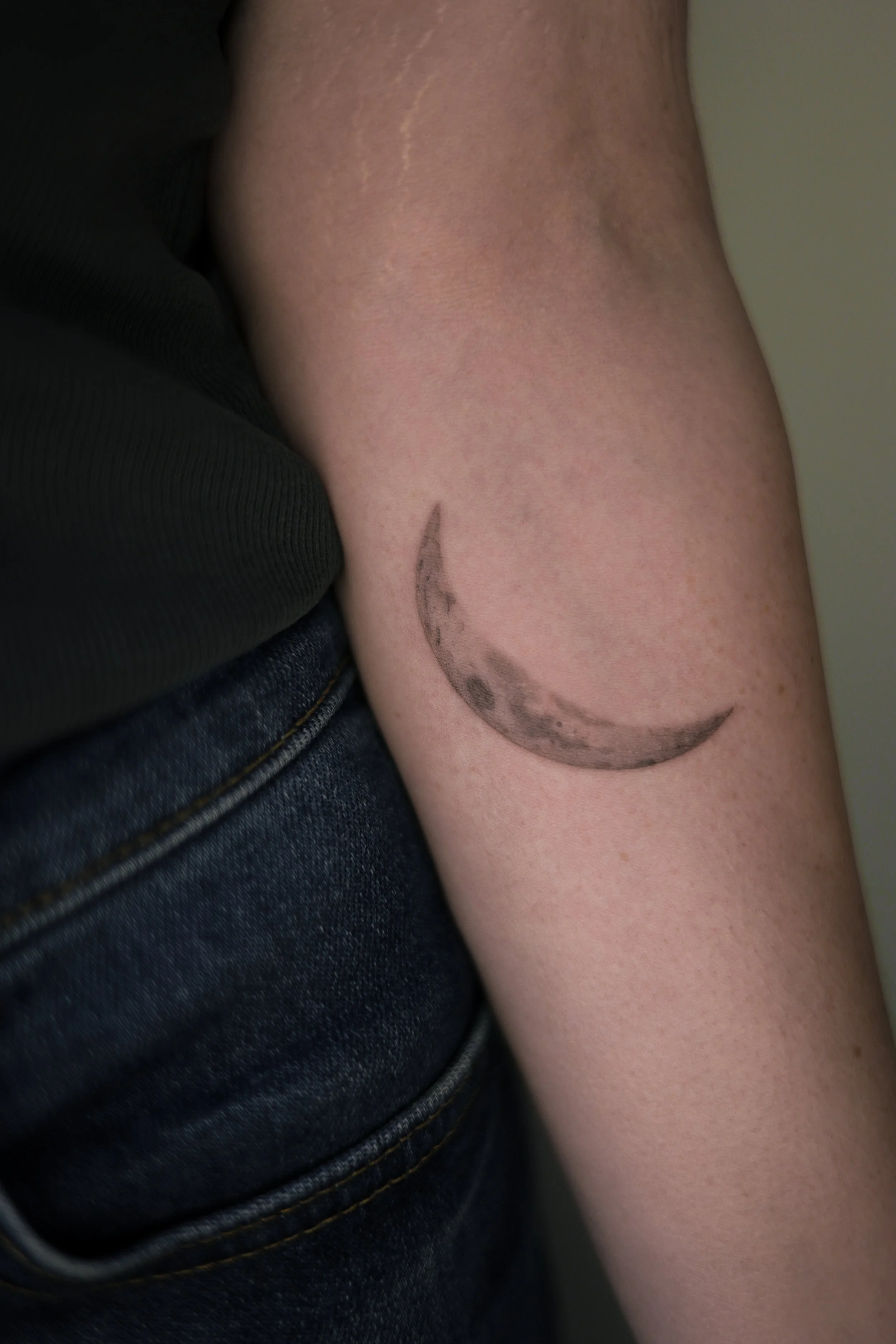 Micro realism crescent moon tattoo on the inner forearm, with detailed lunar texture and soft black and grey shading, by a Vienna tattoo artist.