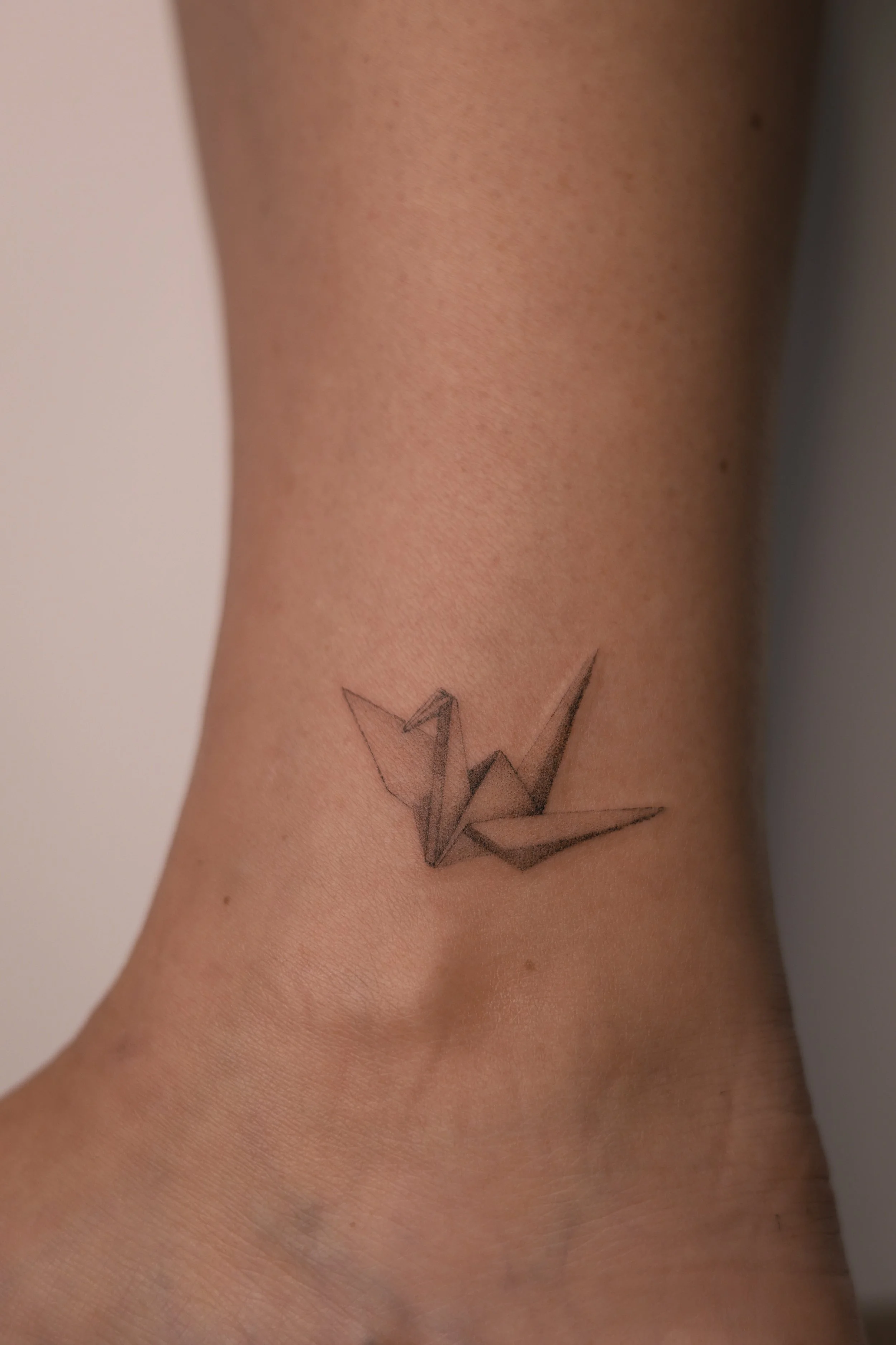 Small micro realism origami crane tattoo on the ankle, with fine lines and soft black and grey shading, by a Vienna tattoo artist.
