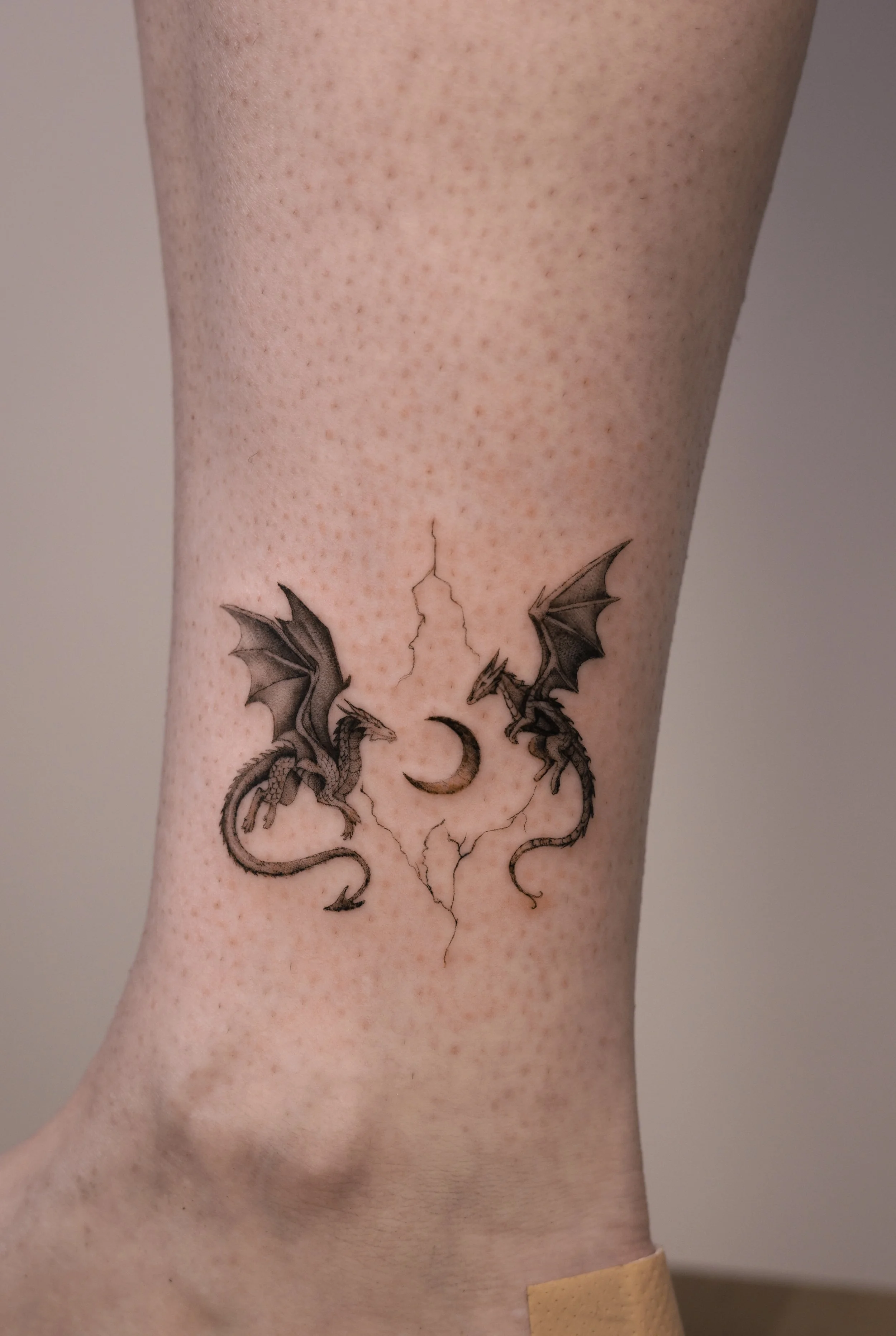 Fine line micro realism dragon tattoo featuring two detailed dragons circling a crescent moon with subtle lightning accents, black and grey fantasy tattoo by Vienna tattoo artist