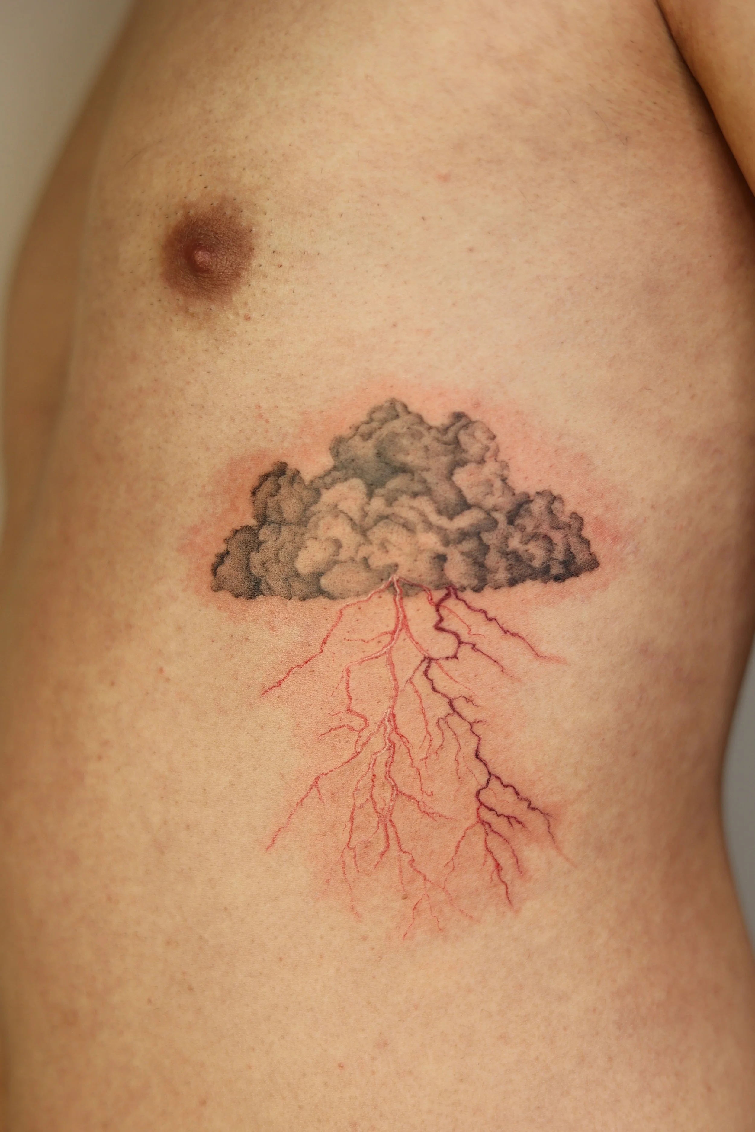 Micro realism storm cloud tattoo on the ribs, with soft black and grey clouds and branching red lightning, by a Vienna tattoo artist.