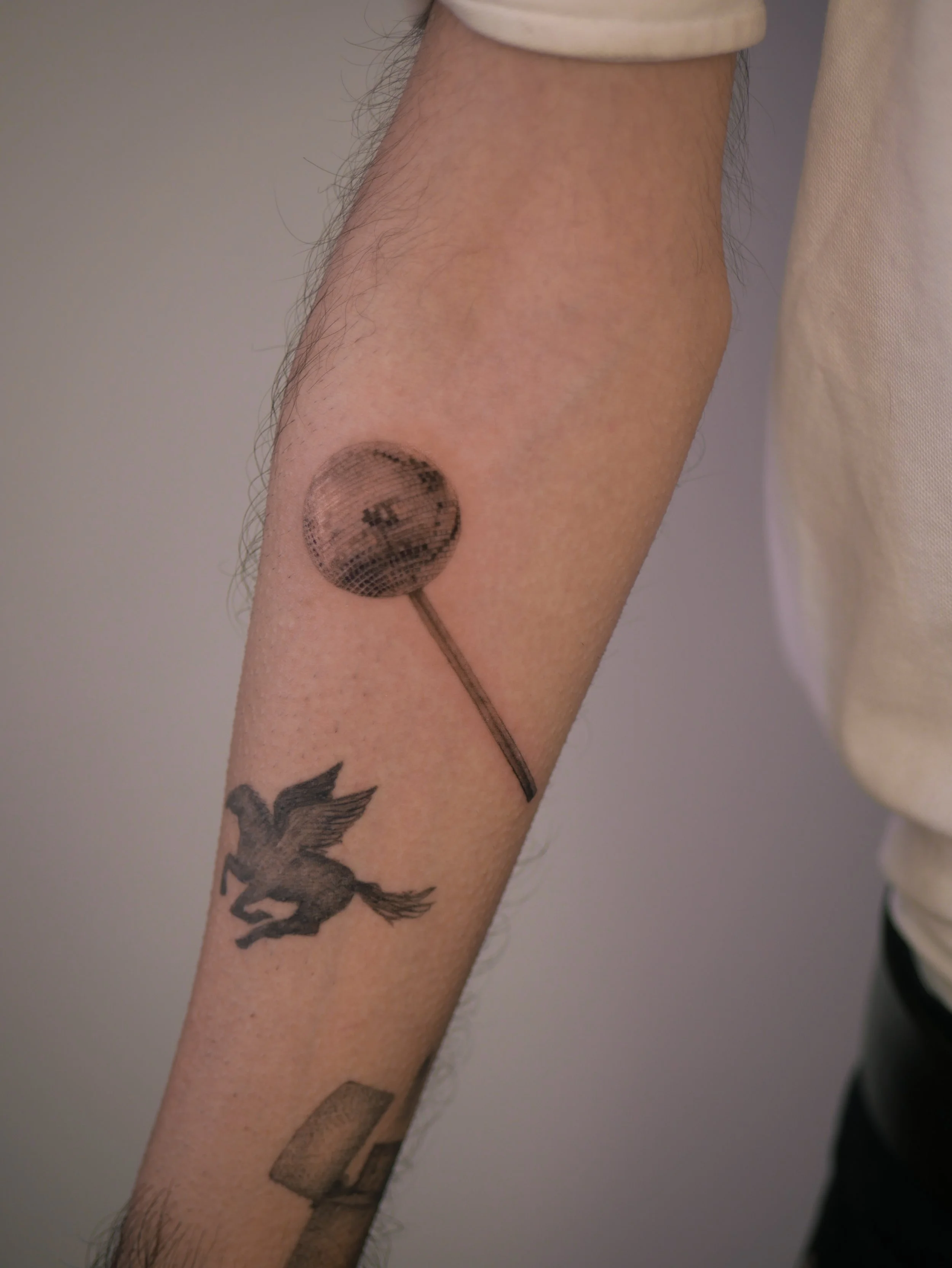 Delicate fine line disco ball lollipop tattoo in black and grey micro realism style on forearm, created by Vienna tattoo artist