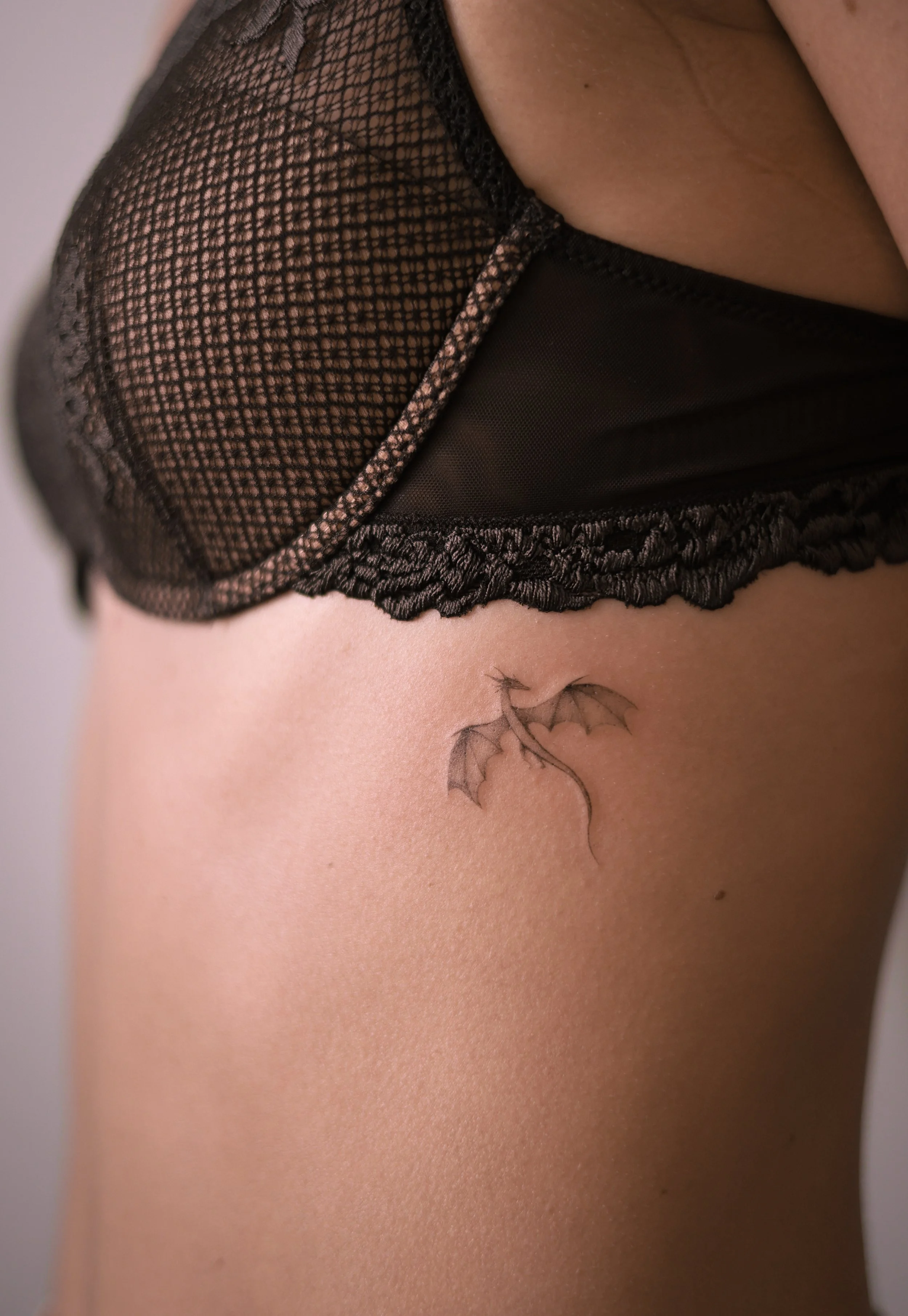 Small dragon tattoo on the ribs, with spread wings, a curved tail and micro realism shading, by a Vienna tattoo artist.