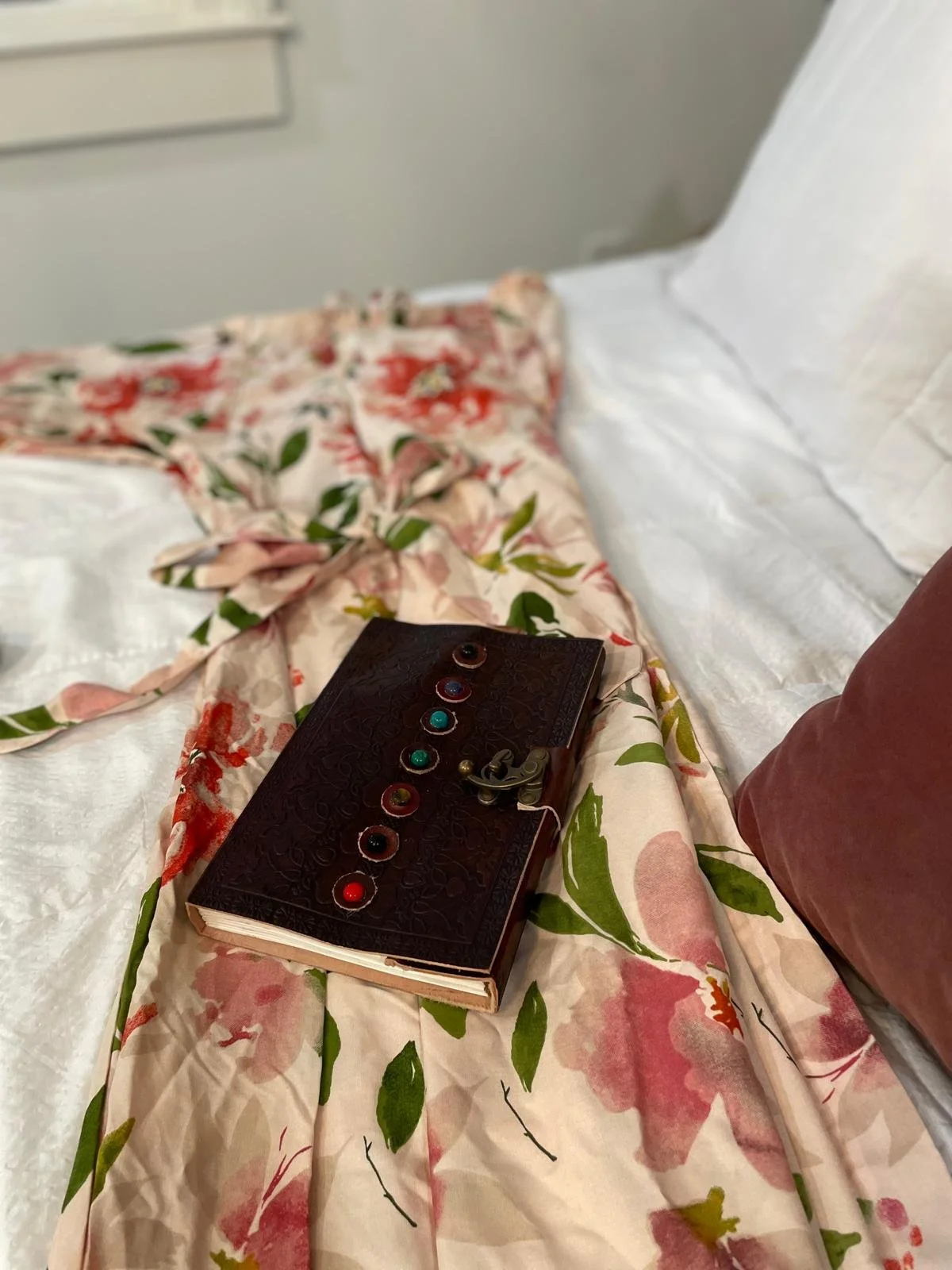 A floral-patterned bedsheet on a bed with a dark brown leather notebook, decorated with multicolored beads, and a small brass lock, adjacent to a person's clothed leg.