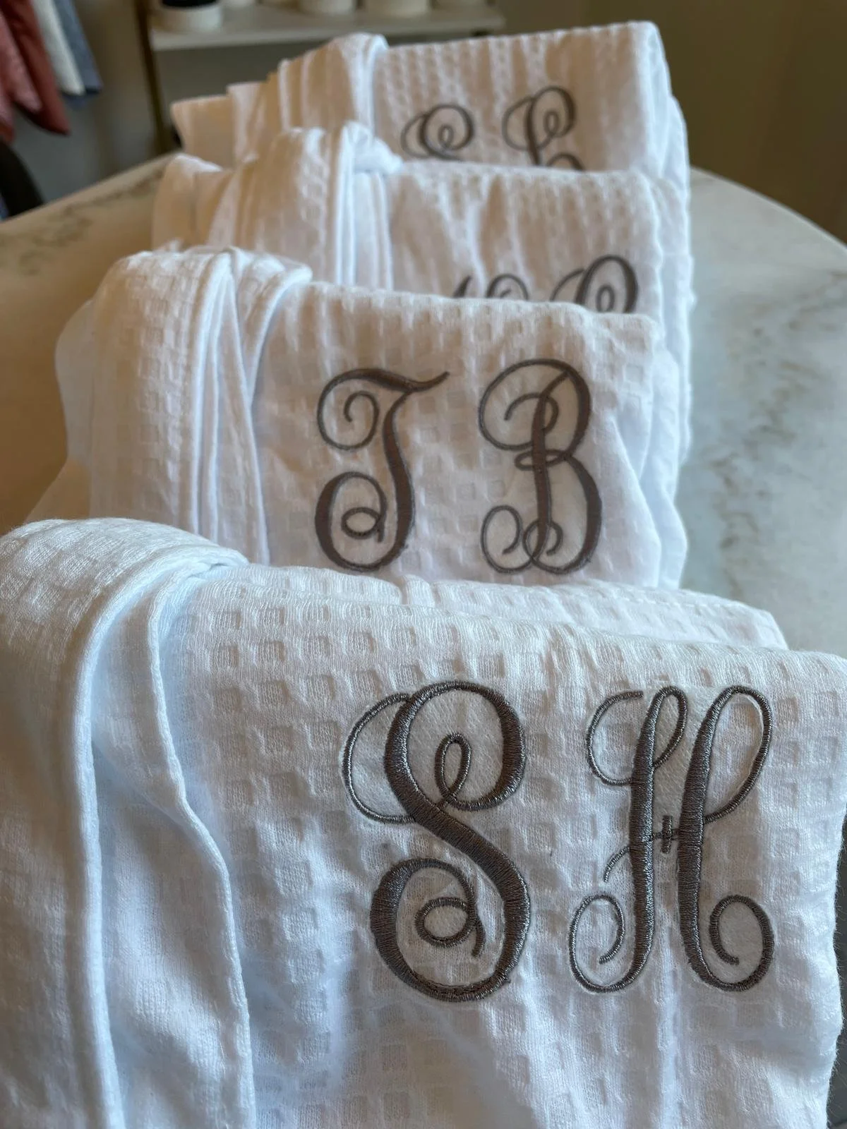 A row of white fabric items with embroidered initials 'T B' in brownish-gray thread, draped over a surface.