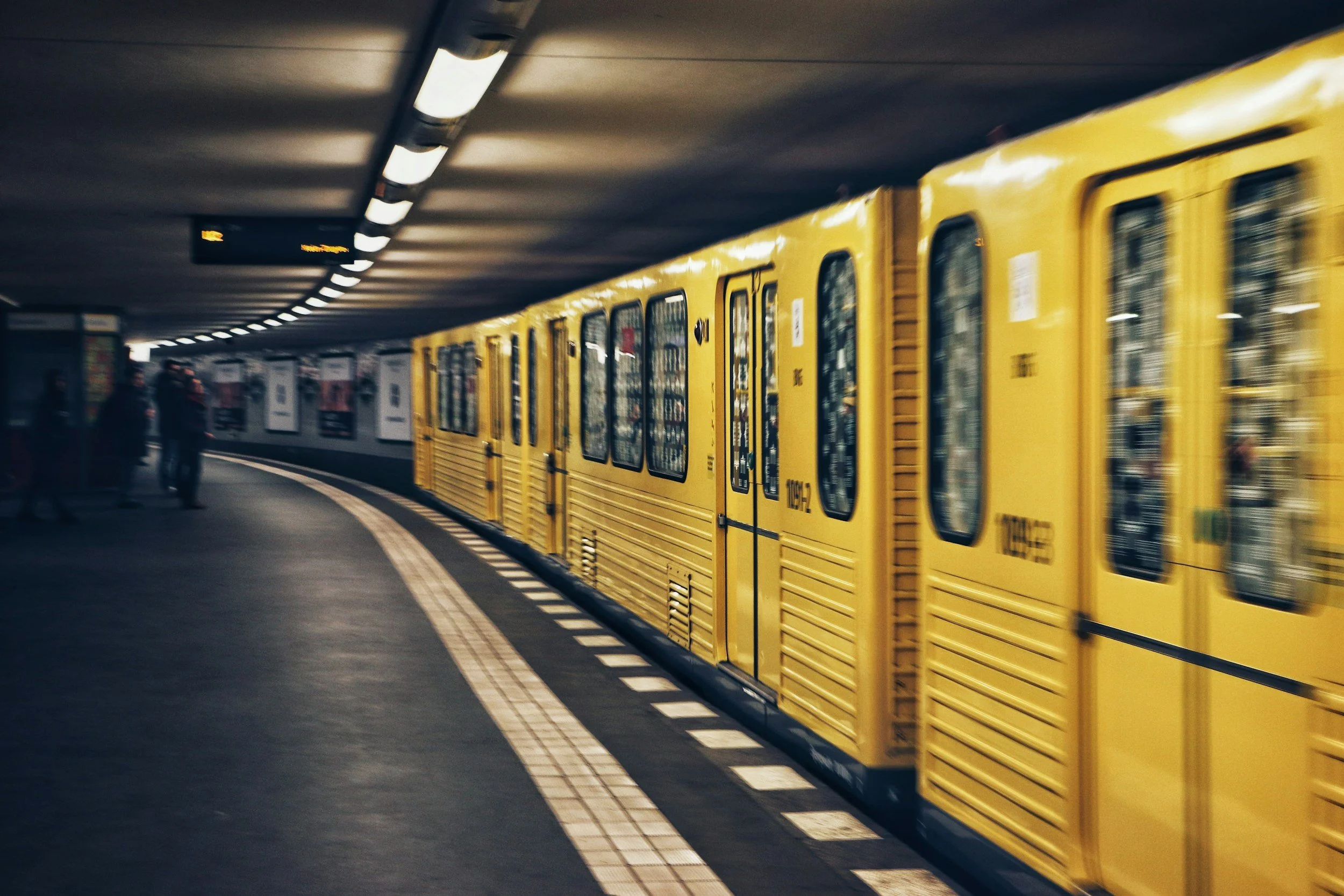 Yellow subway train stopped in an underground station with a curved platform and pedestrians nearby.