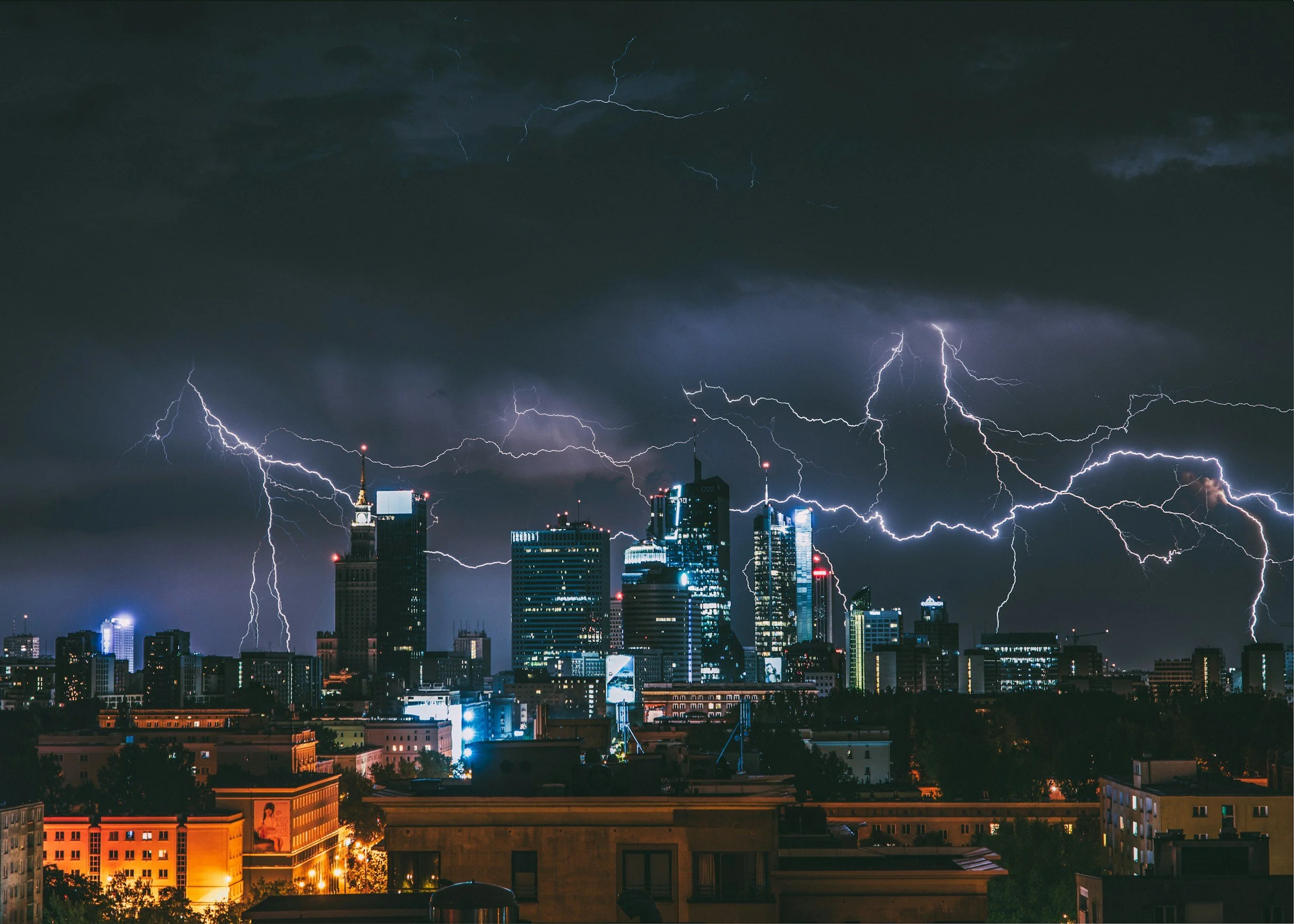 Nighttime cityscape with multiple skyscrapers illuminated against dark stormy sky, featuring prominent lightning strikes striking the buildings.