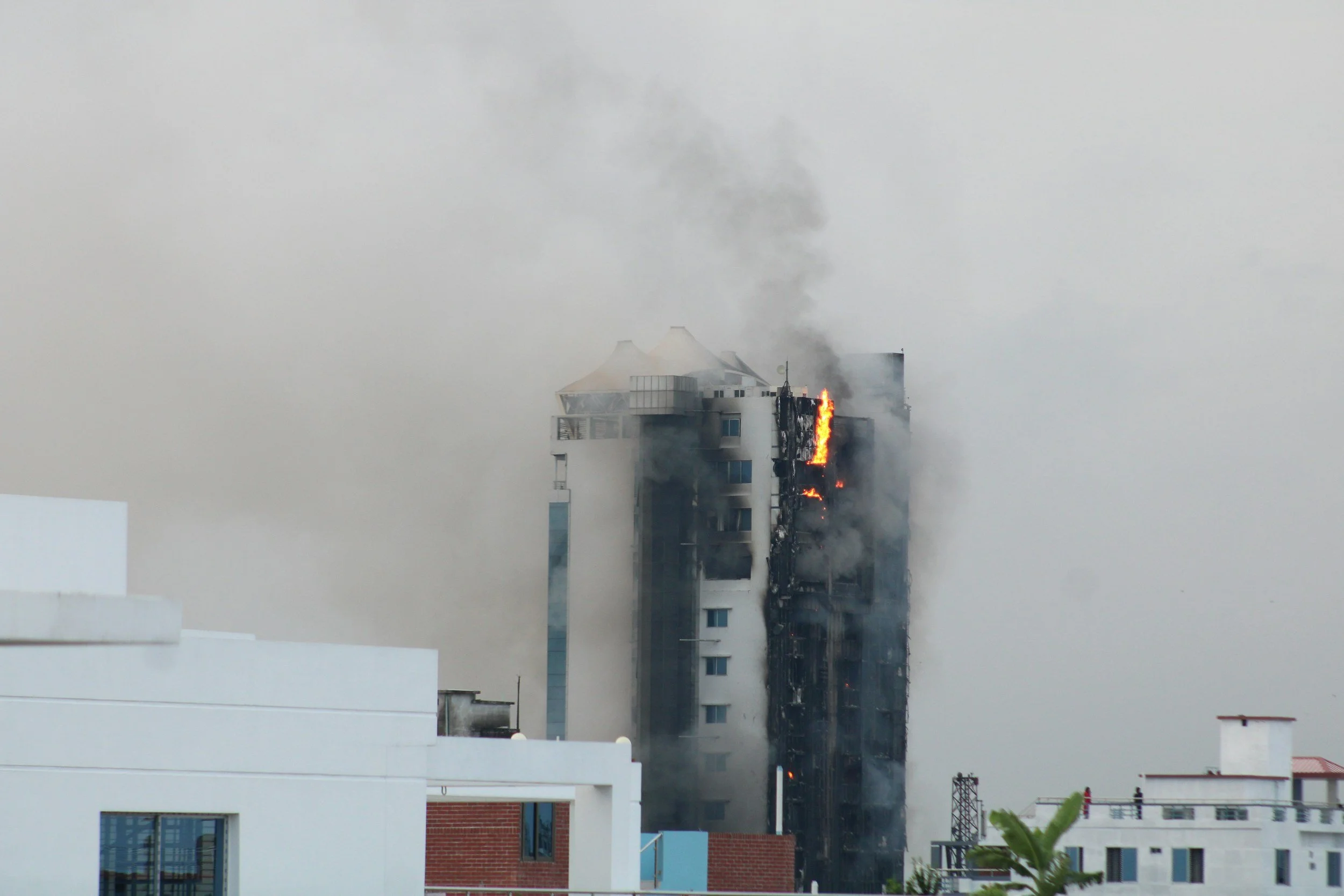 A multi-story building on fire with black smoke and flames visible from a window on one floor, surrounded by gray overcast sky.