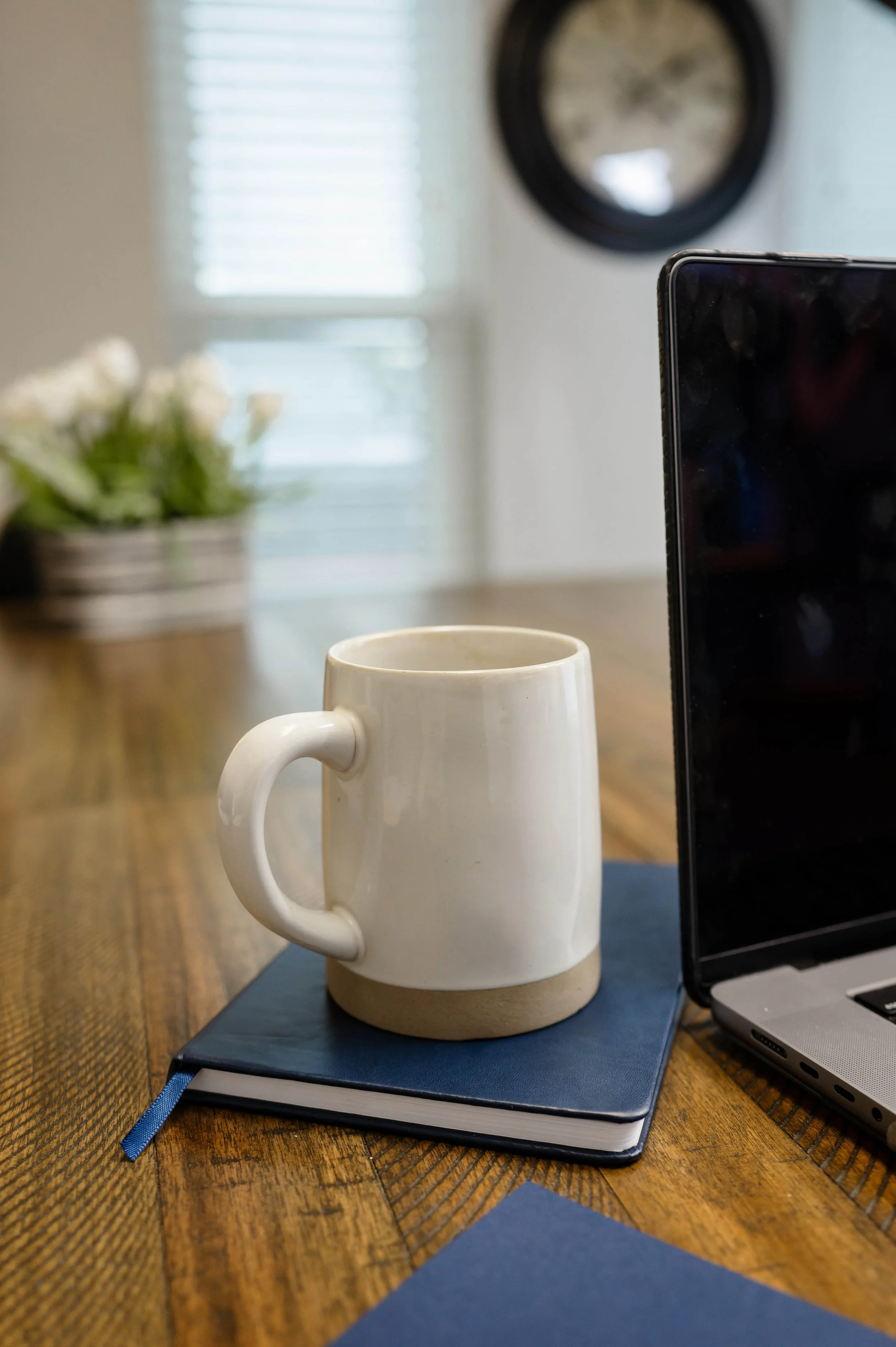 A white ceramic mug placed on a closed blue notebook on a wooden table with a laptop to the right. In the background, a blurry potted plant and a round wall clock are visible.