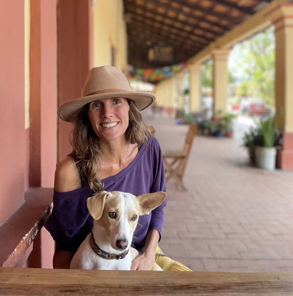 A woman with shoulder-length wavy hair wearing a beige wide-brimmed hat and a purple top, smiling while sitting at an outdoor table with a tan and white dog with large ears in her lap. The background shows a covered patio with potted plants, colorful decorations, and a brick walkway.