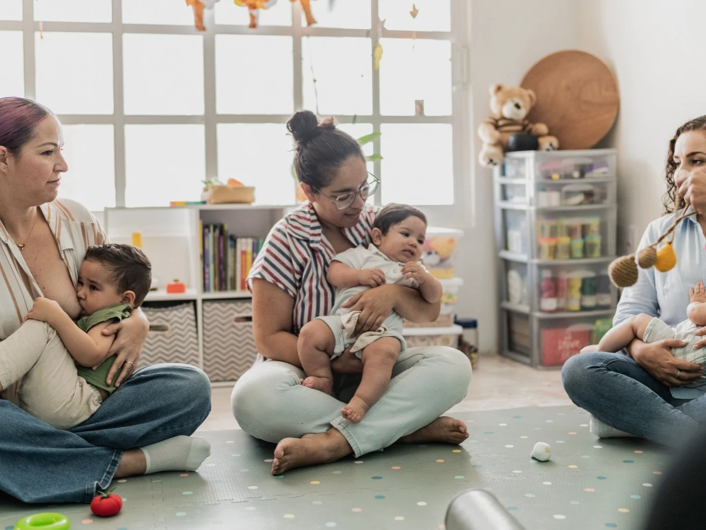 Three women sitting cross-legged on the floor in a bright room, each holding a small child. The women are engaged in a group activity with the children, with toys scattered on the mat. Background includes a window with sunlight, shelves with toys and books, and a teddy bear on a cabinet.
