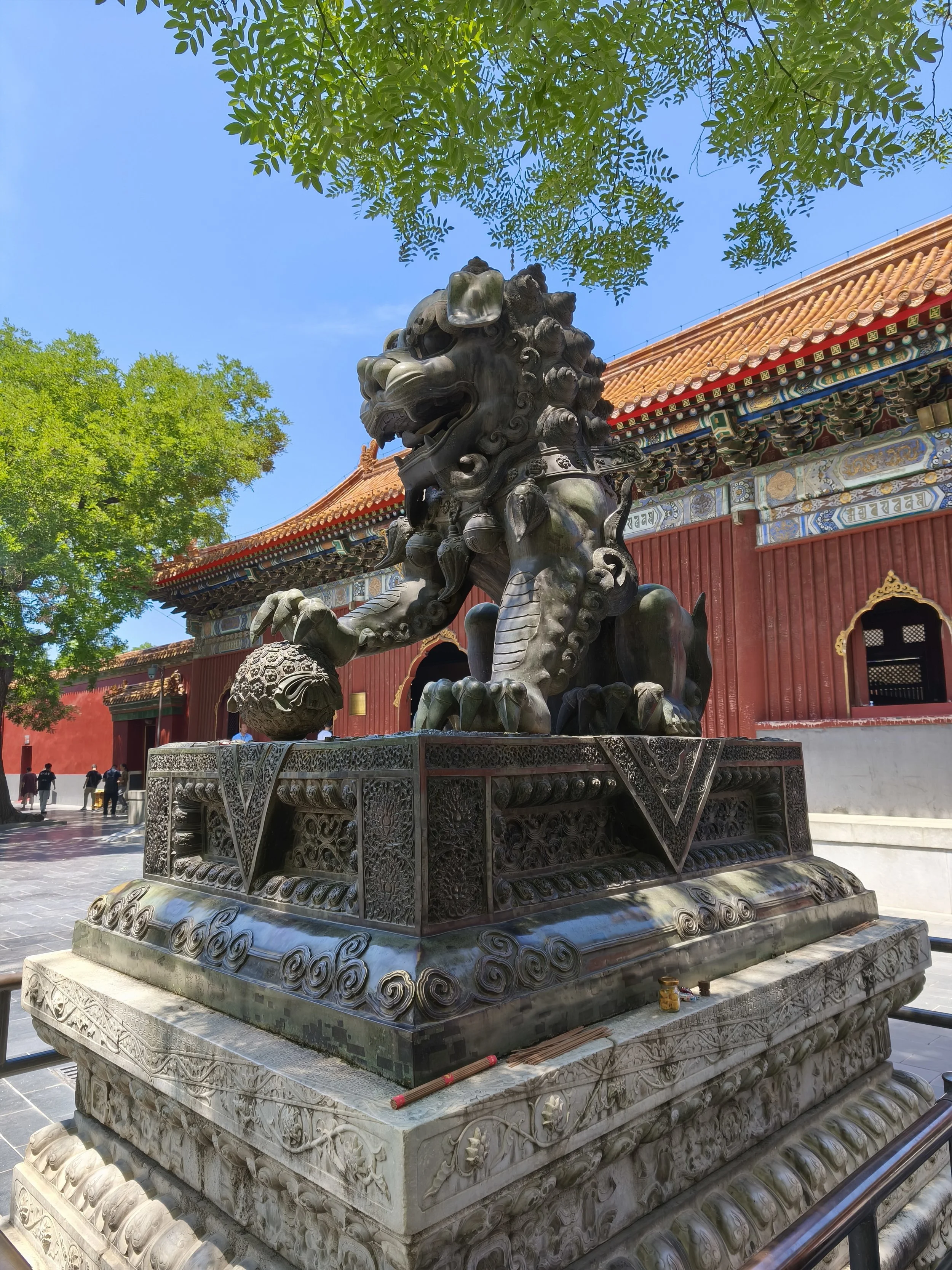 A large bronze Chinese guardian lion statue in front of a traditional Chinese building with red walls and intricate colorful roof decorations, surrounded by trees with green leaves and a blue sky.