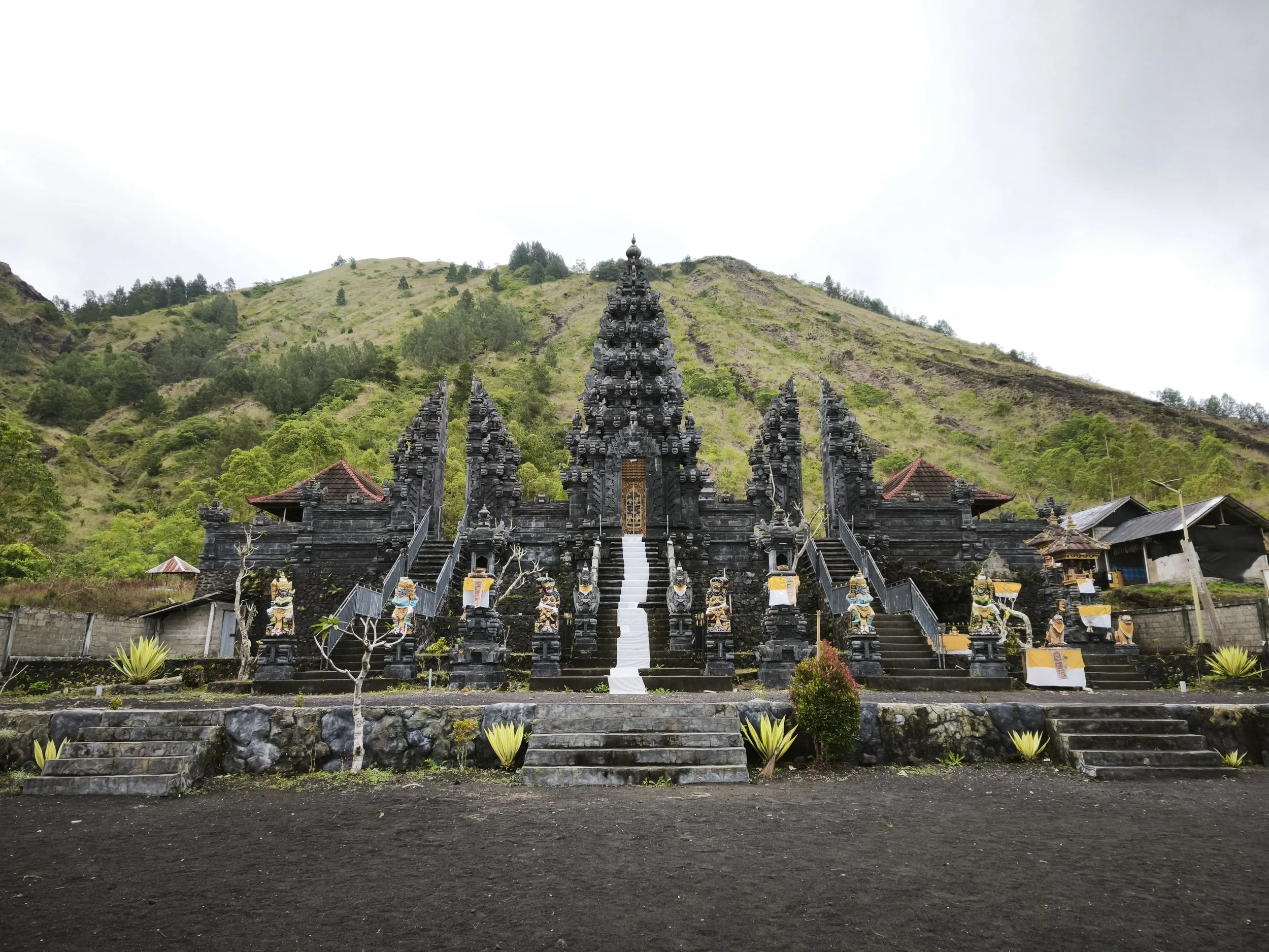 A traditional Balinese temple made of dark stone, with intricately carved staircases and statues, set against lush green hills under an overcast sky.