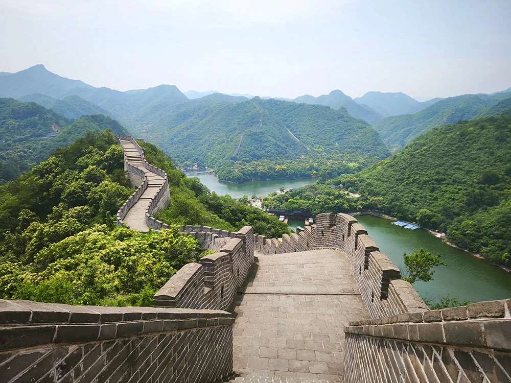 Kilátás a kínai nagy falra, amely zöld hegyeken és alatta egy folyón kanyarog buja, hegyvidéki tájban.