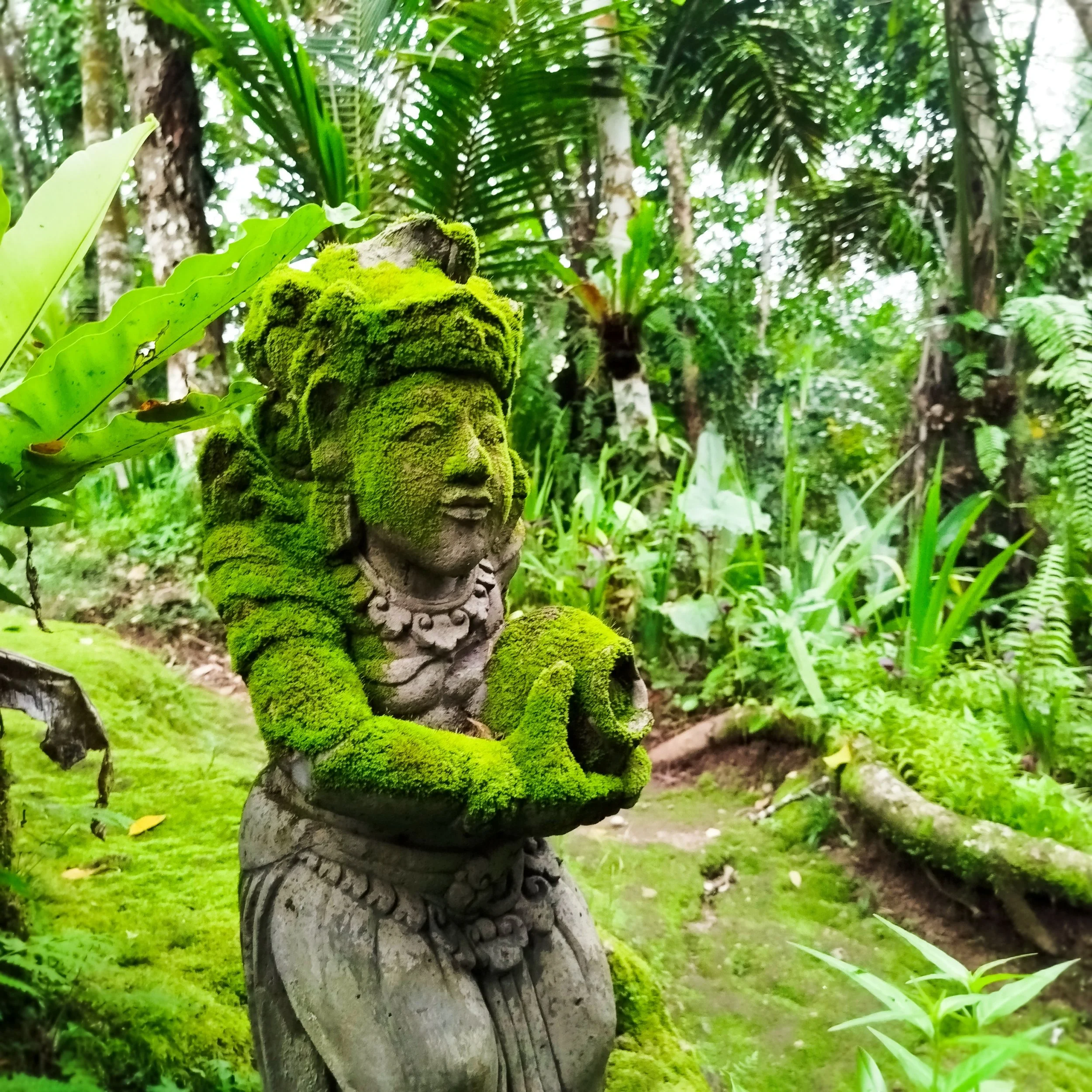 A moss-covered stone statue of a woman with intricate jewelry holding an object, situated in a lush green tropical forest.
