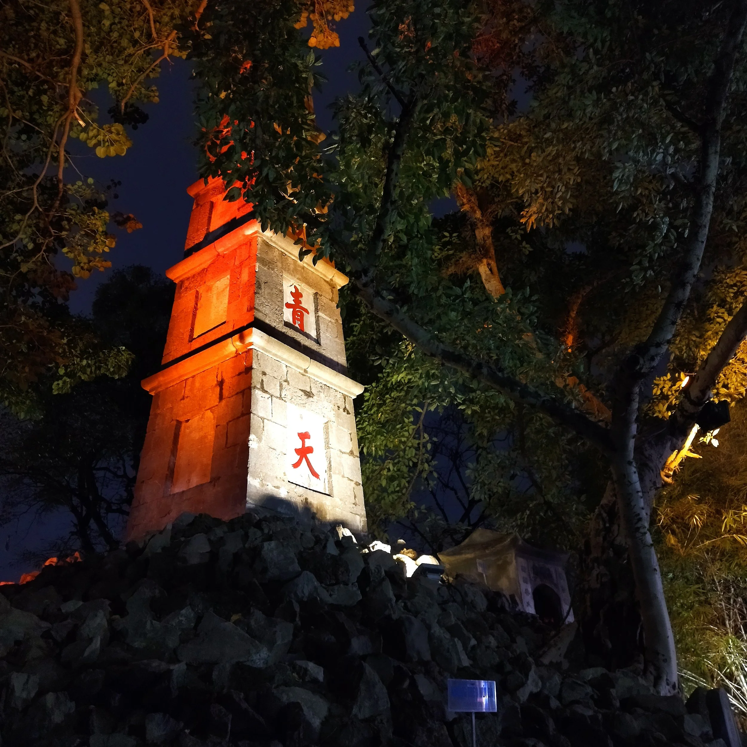 A tall stone tower illuminated with orange and white lights at night, with Chinese characters in red on white plaques on its front, surrounded by trees and rocks.