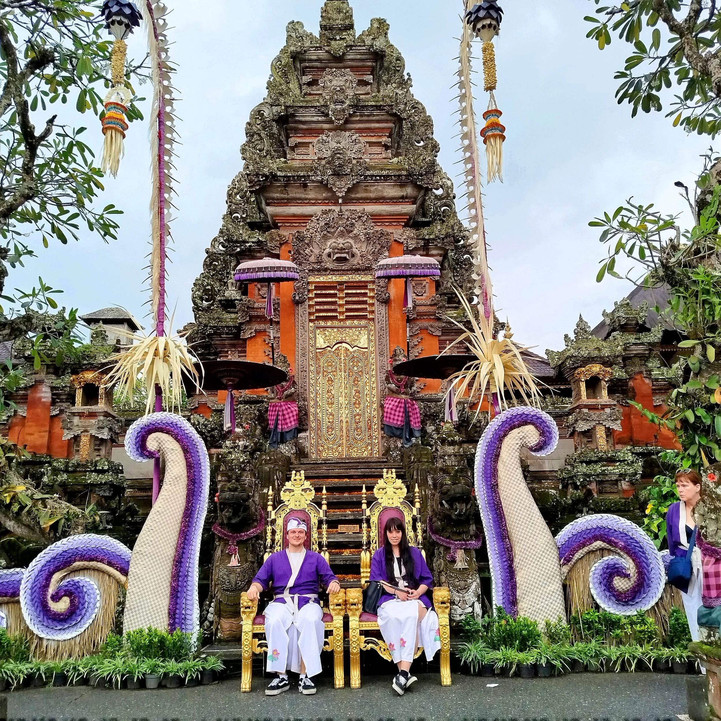 A traditional Balinese temple with ornate carvings and a gold gate. In front, two people in purple and white staff uniforms sit on gold chairs. Decorated mythical serpent statues and offerings surround them, with decorations hanging from trees.
