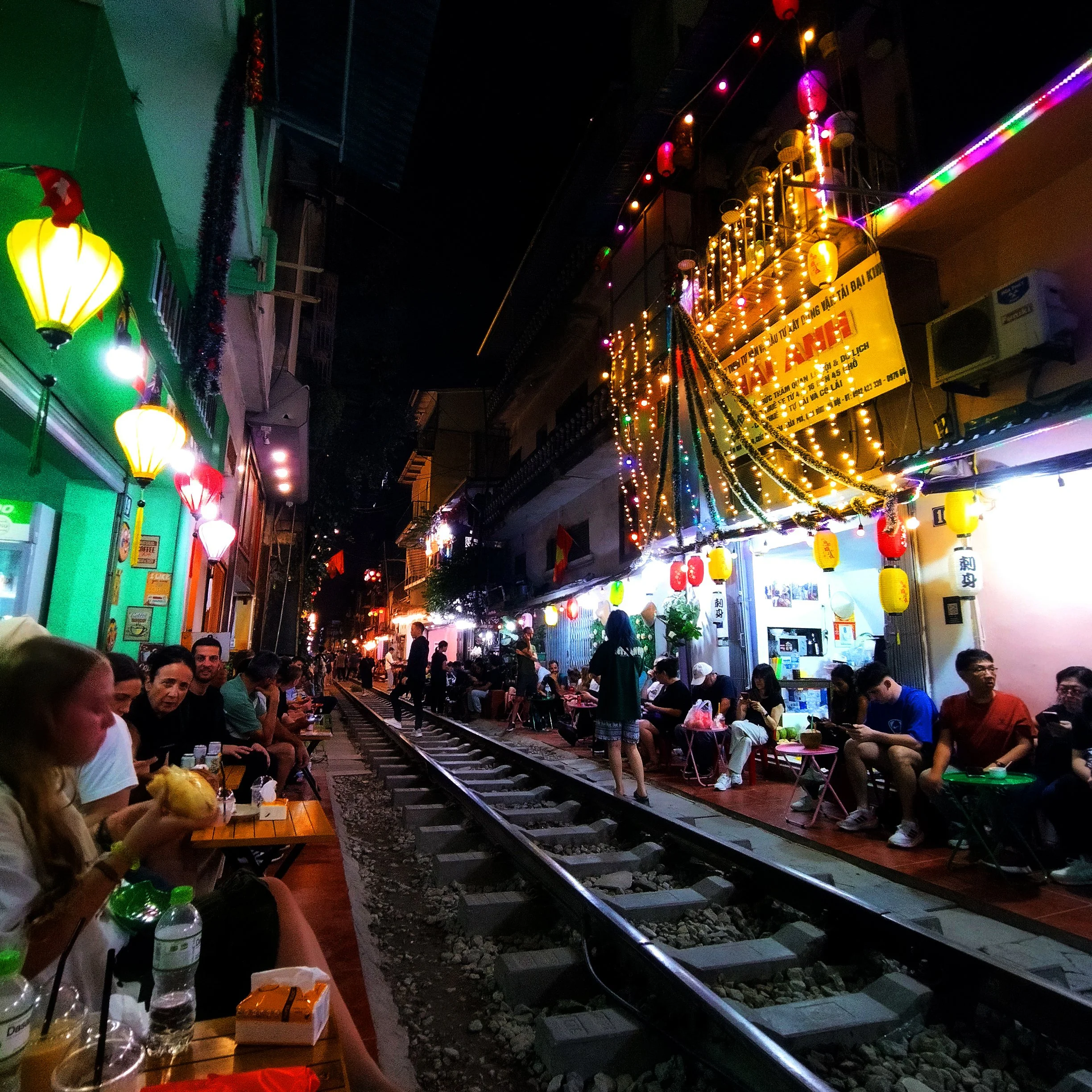 Night scene of a street festival with people sitting along the railway tracks, lit with colorful lanterns and string lights, with shops and restaurants on either side.