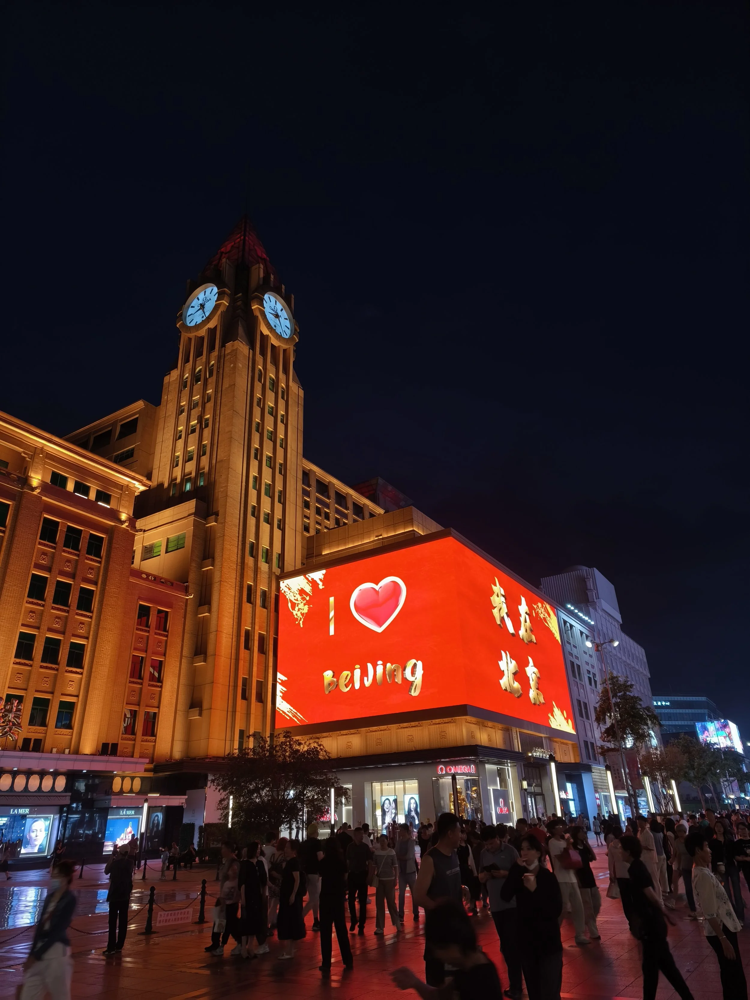 Night view of a busy city square in Beijing with illuminated buildings, a large electronic billboard displaying a red background with a heart symbol and the words "I love Beijing" in English and Chinese, and a clock tower showing the time as 9:20, with many people walking around.
