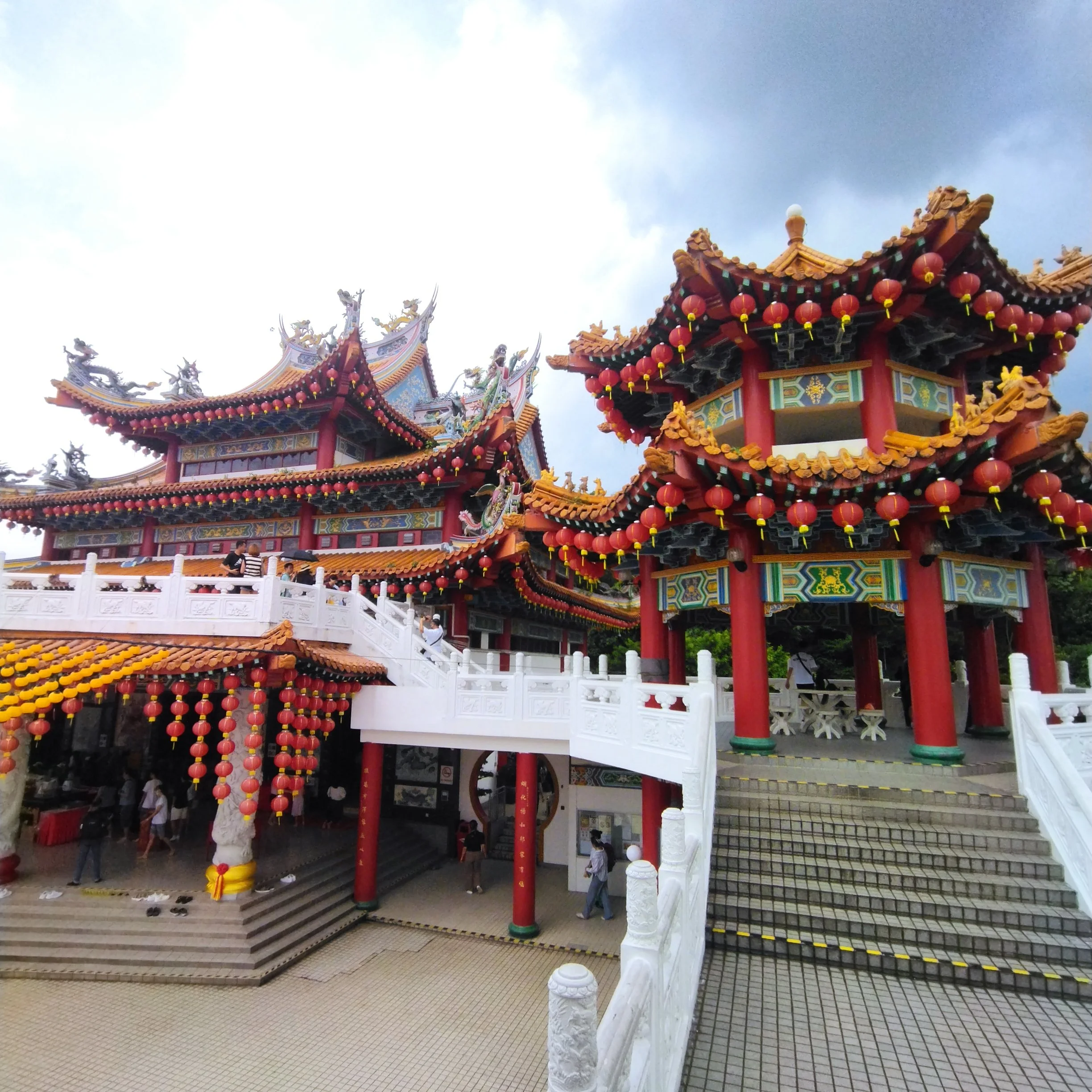 A traditional Chinese temple with ornate, colorful roofing decorated with dragons and lanterns, surrounded by a white stone railing, under a cloudy sky.