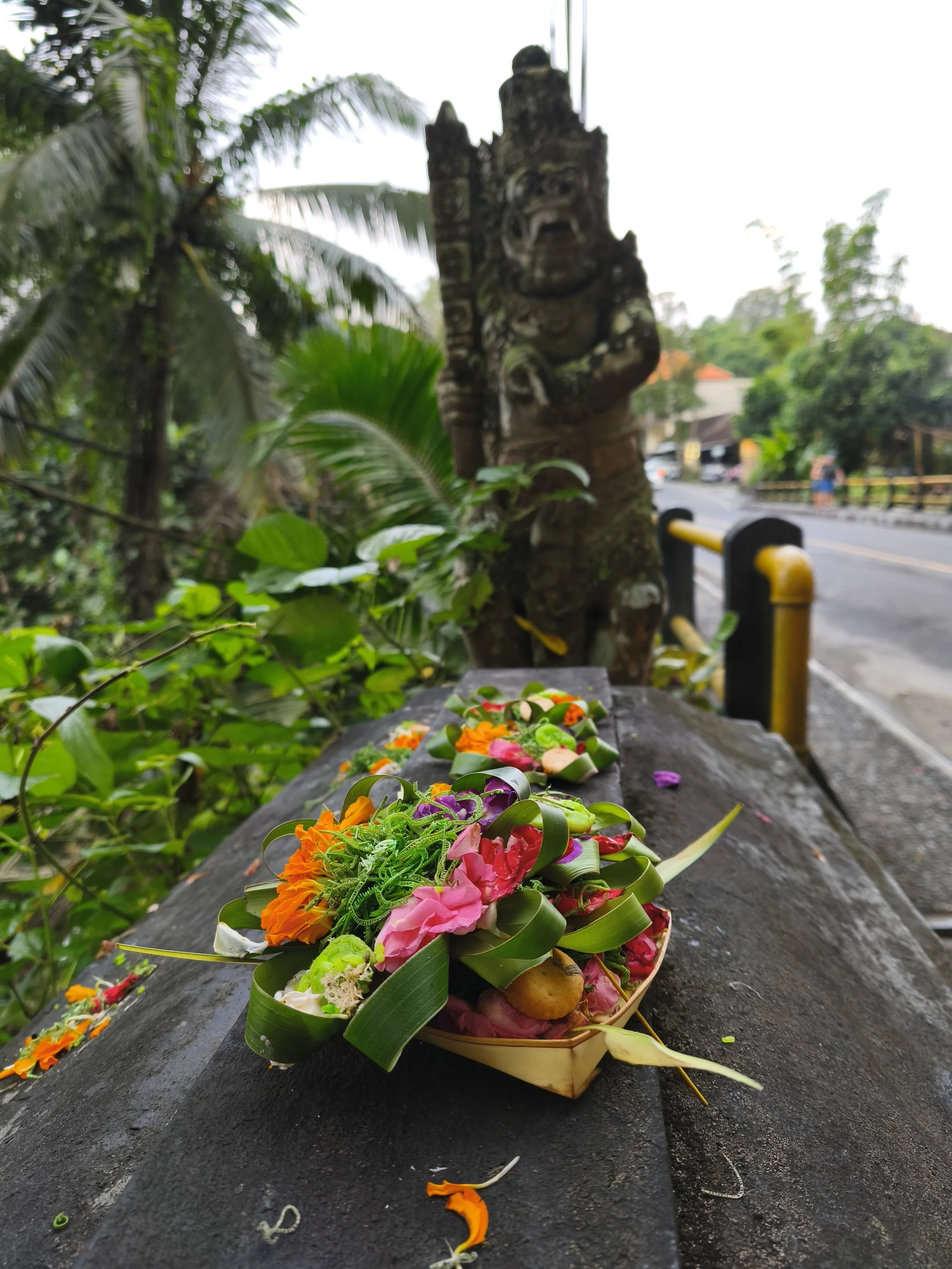 Színes virágokkal és levelekkel díszített Balinéz áldozat, kőfelületre helyezve, egy vad arcú, faragott kőszobor mellett, buja zöld kültéri környezetben. Balin