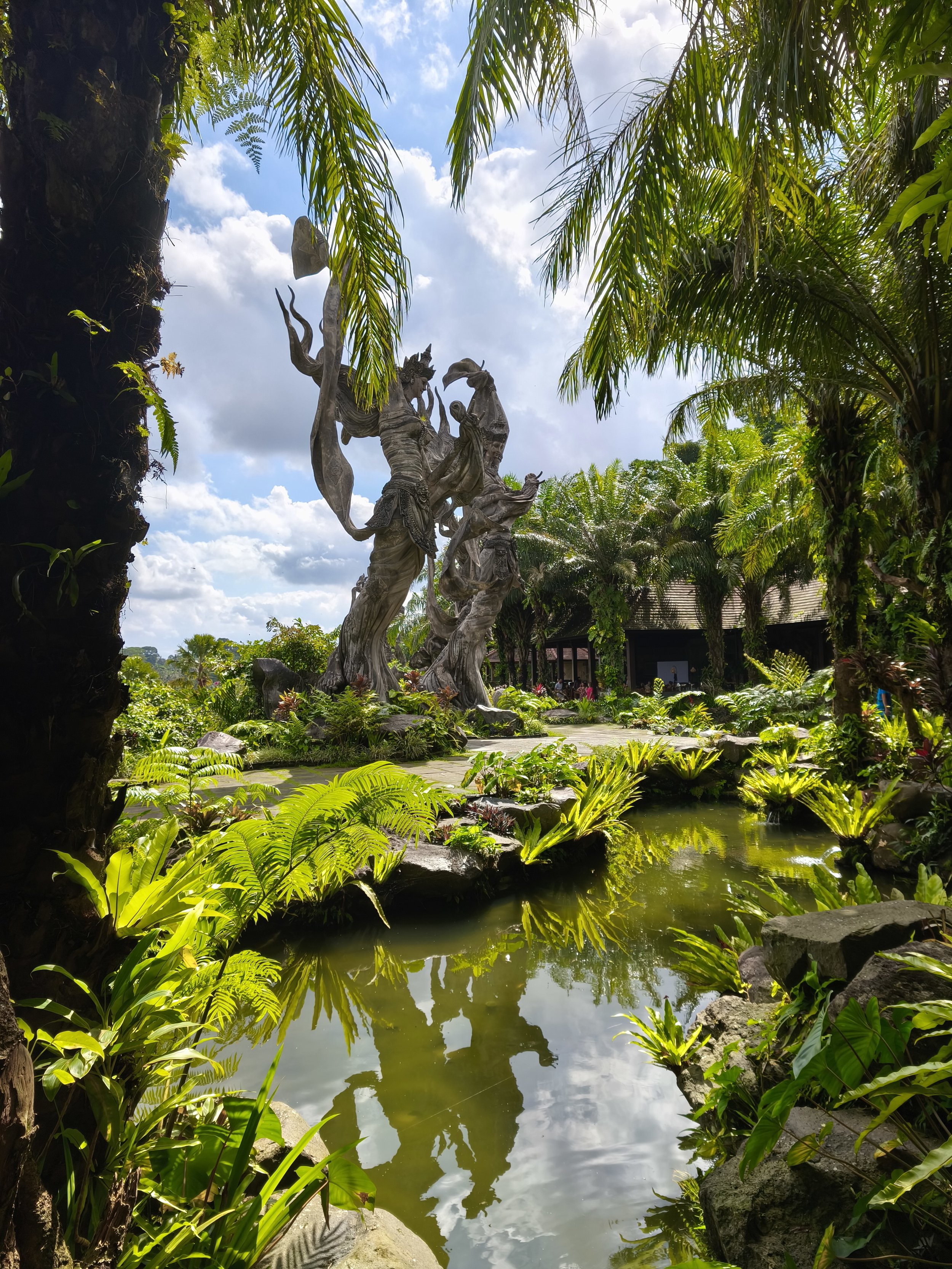 A tropical garden with lush green plants, a pond reflecting the sky, and a large wooden sculpture of mythological figures.