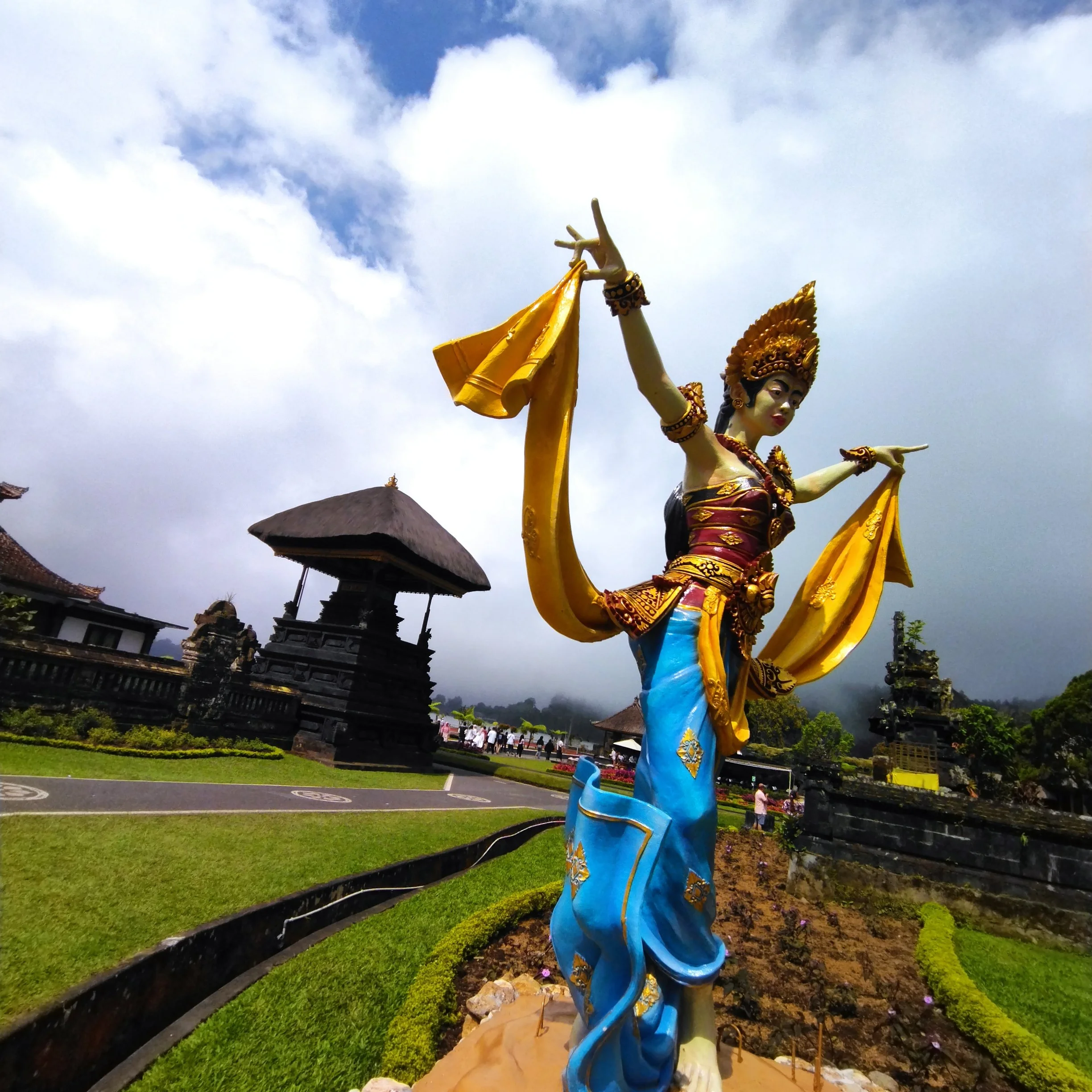 Colorful sculpture of a woman in traditional attire with outstretched arms, standing in a landscaped garden with traditional buildings and cloudy sky in the background.