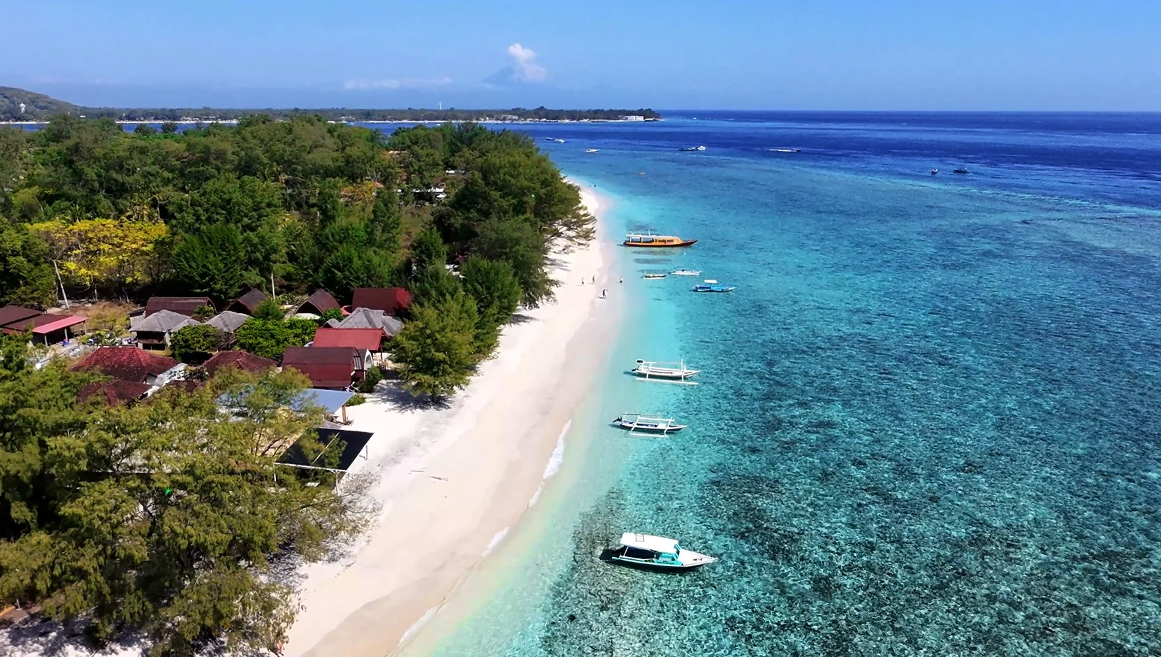 Aerial view of a tropical beach with white sand, clear turquoise water, and boats docked along the shore. Green trees and small houses line the coast, with a distant landmass visible in the background under a partly cloudy sky.