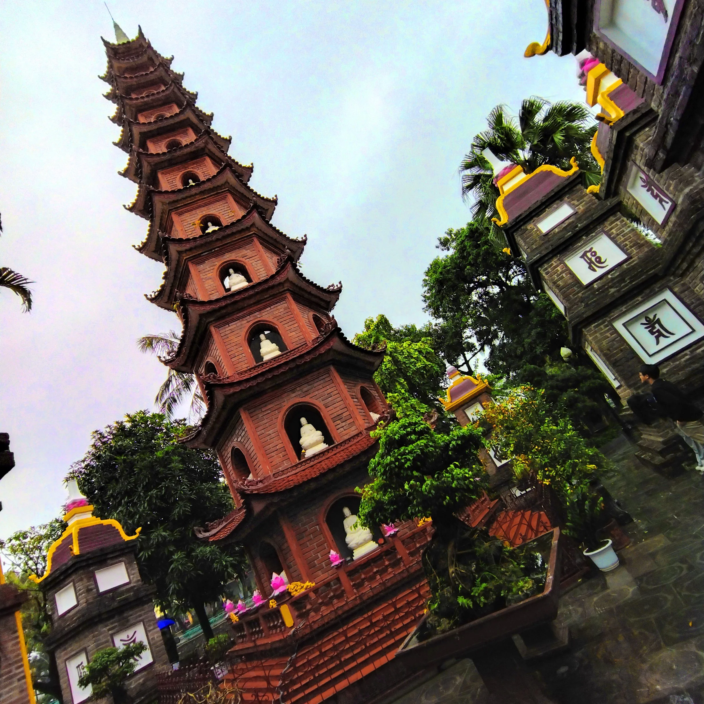 A multi-tiered, red pagoda with small white Buddha statues on each level, surrounded by plants and trees, with decorative brick walls and a cloudy sky in the background.