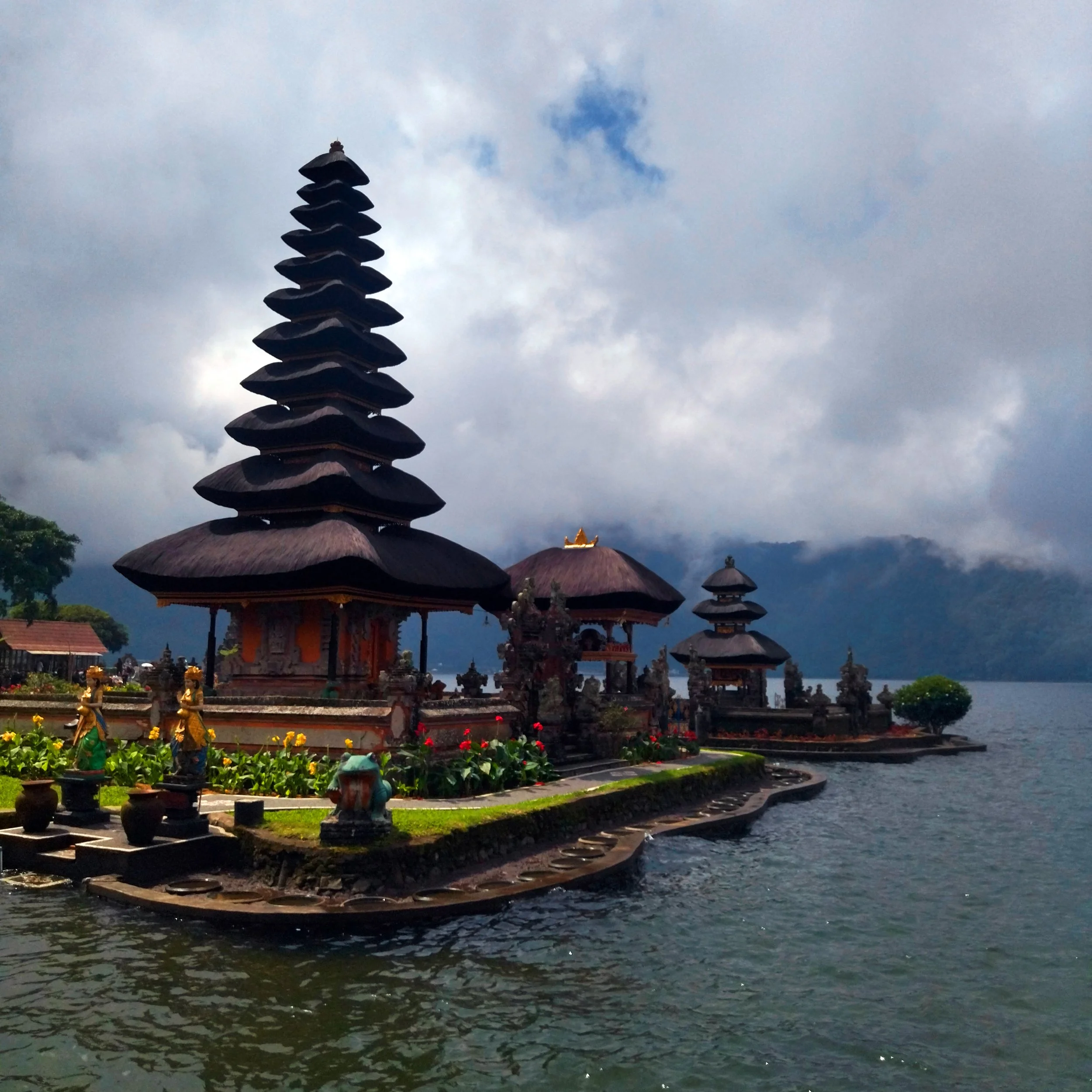 Traditional Balinese temple structures with multiple tiered thatched roofs on a small island surrounded by water, with cloudy sky and mountains in the background.