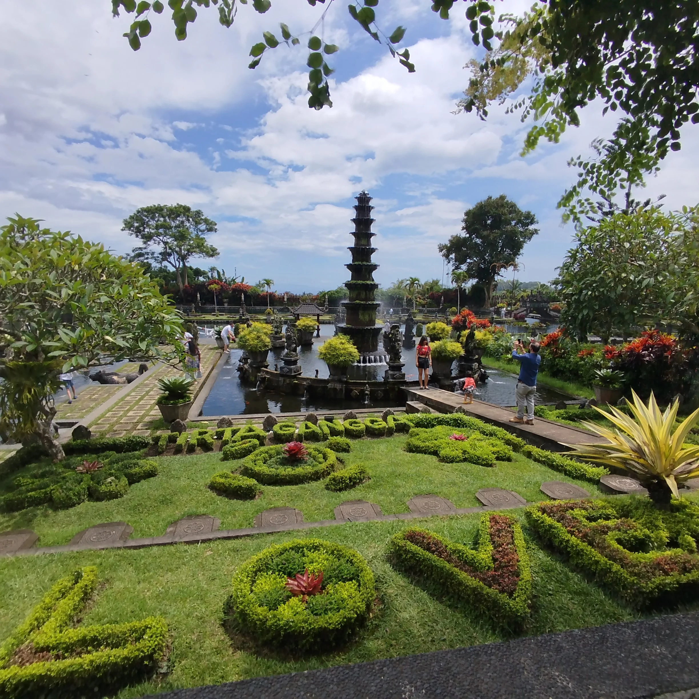A lush garden scene at Tirta Gangga with a central tiered fountain in a pond, surrounded by vibrant flowers, trimmed hedges, and visitors taking photos on a sunny day with a partly cloudy sky.