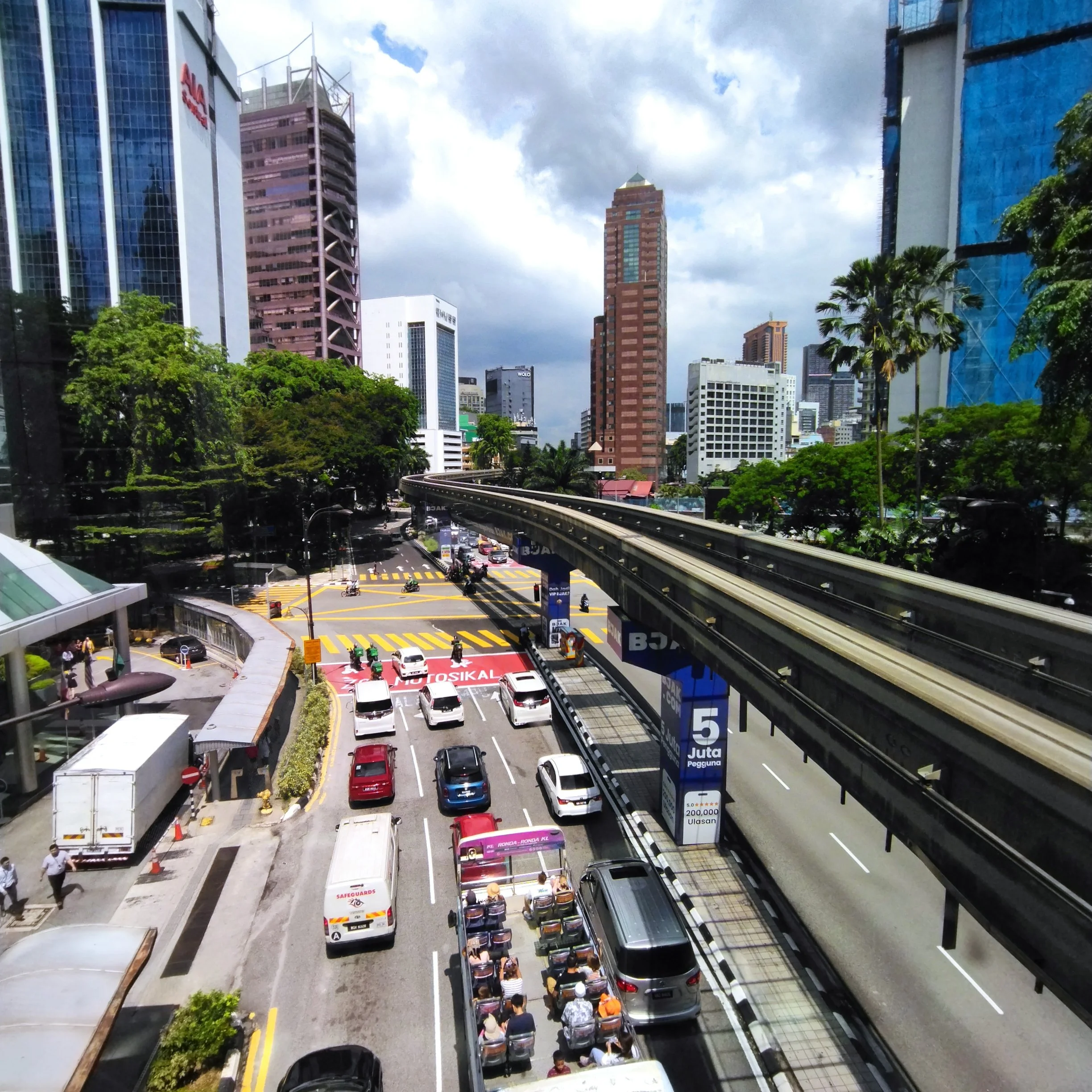 A cityscape with tall buildings, a train track elevated above the street, and a traffic jam with cars, buses, and motorcycles at a crosswalk under a partly cloudy sky.