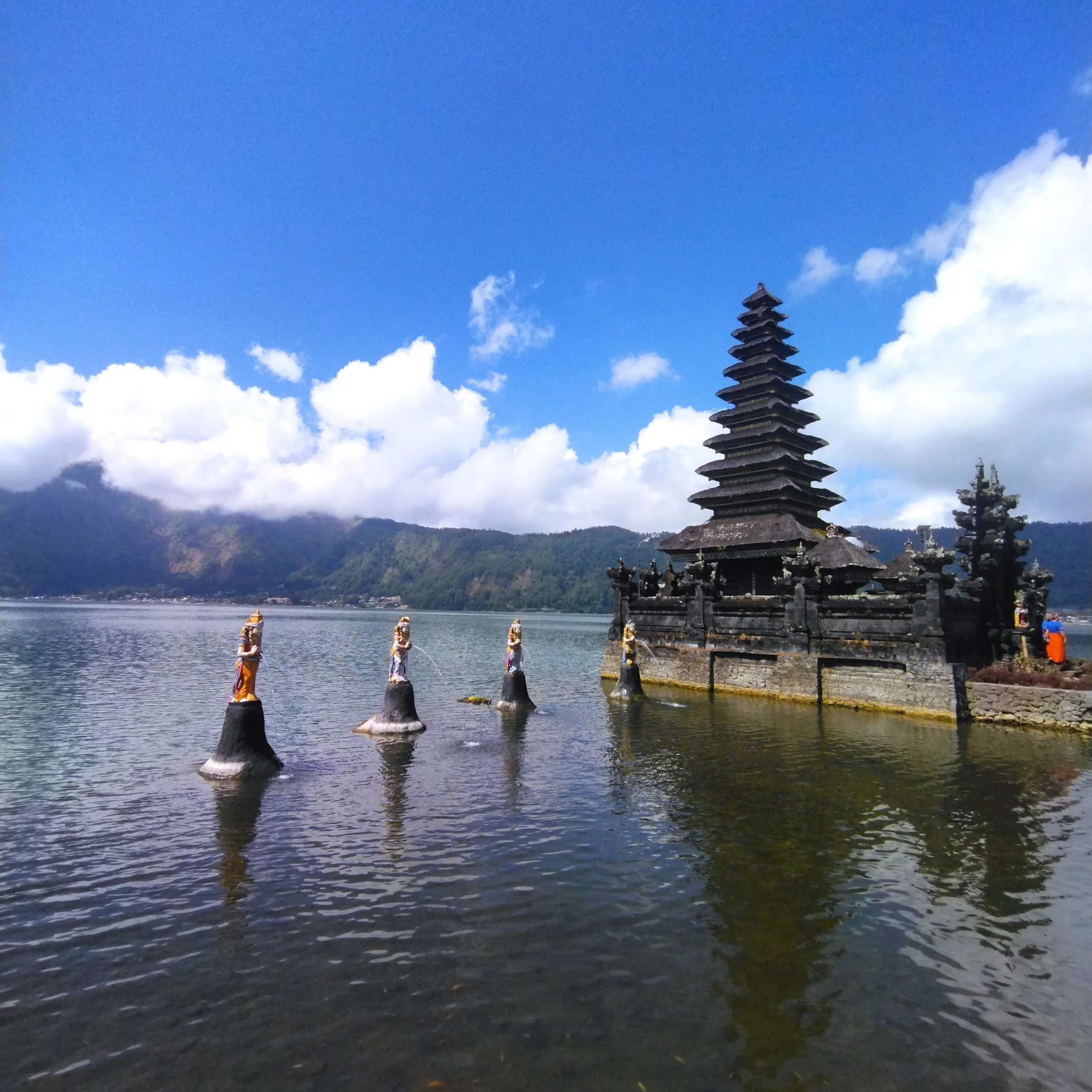 A traditional black pagoda on the shore of a lake with four stone statues of guardian figures with gold details in the water. Mountain range and partly cloudy sky in the background.