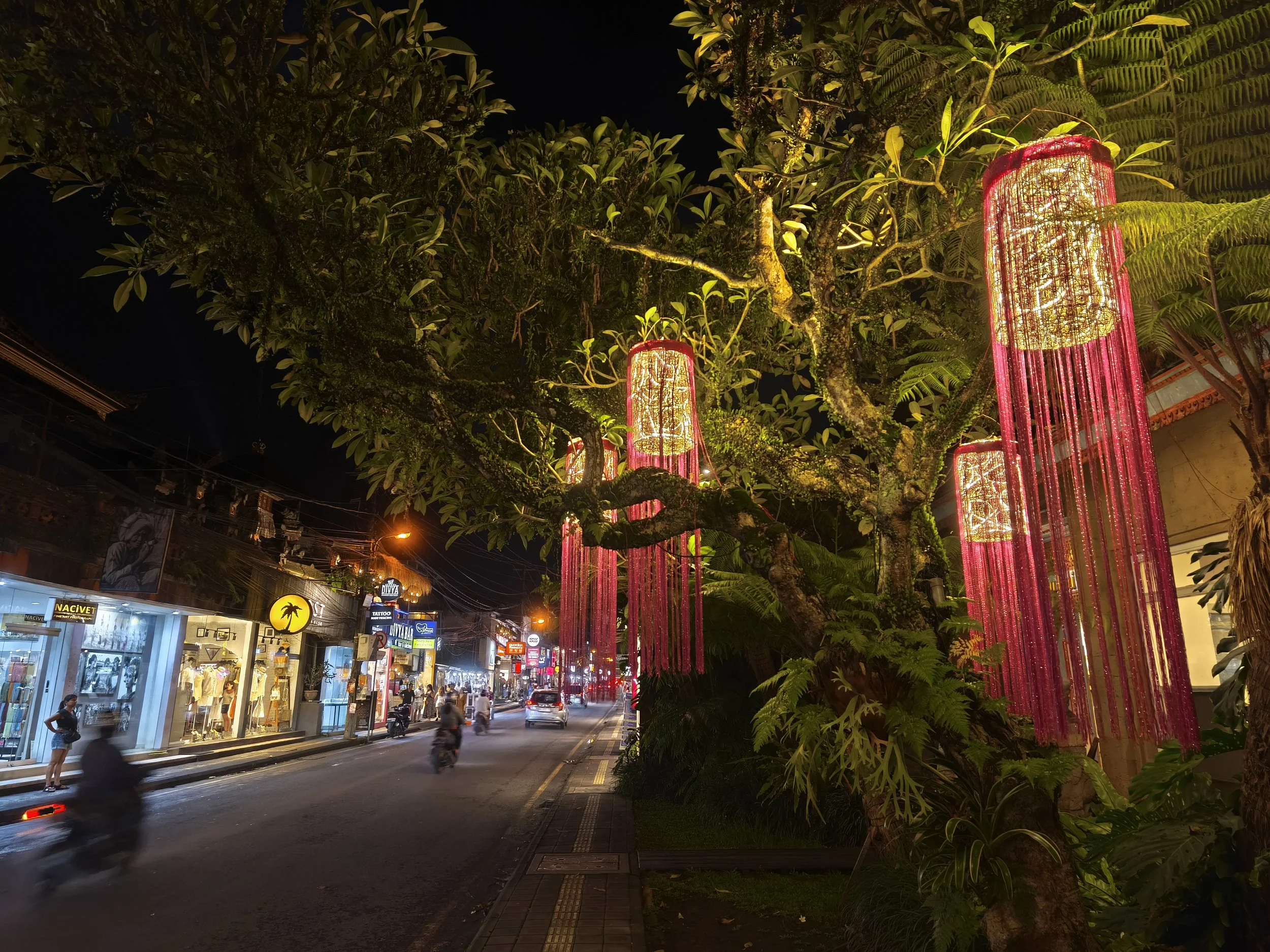 Nighttime street scene with shops on the left, including a shop with a yellow sign and some people walking or riding motorcycles. On the right, there are trees decorated with pink hanging lamp-like ornaments emitting yellow light, creating a festive atmosphere.