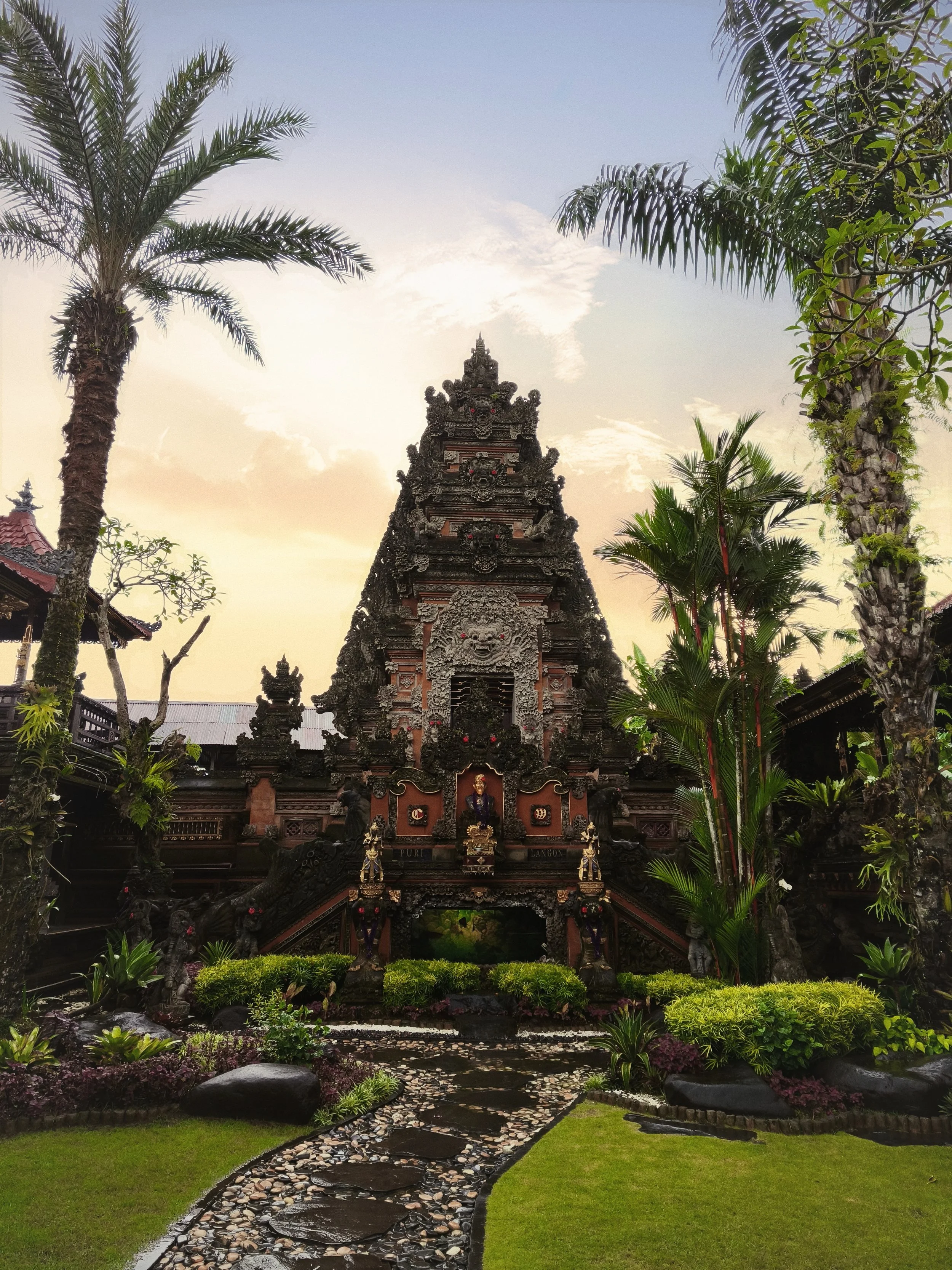 Egy hagyományos balinéz templom bonyolult kőfaragásokkal, buja zöld növényekkel és pálmafákkal körülvéve naplementekor.